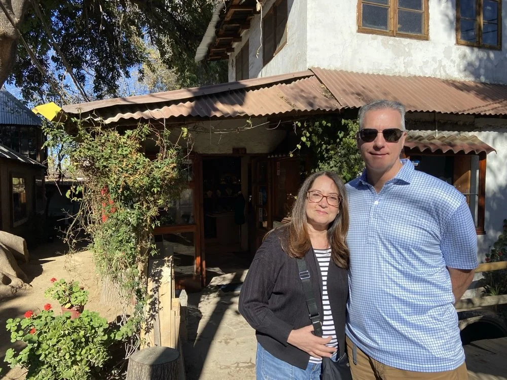 A woman and a man stand together outside a rustic building with a corrugated metal roof, trees, and potted plants. The woman wears glasses and a striped shirt with a black jacket, while the man wears sunglasses and a blue checkered shirt.