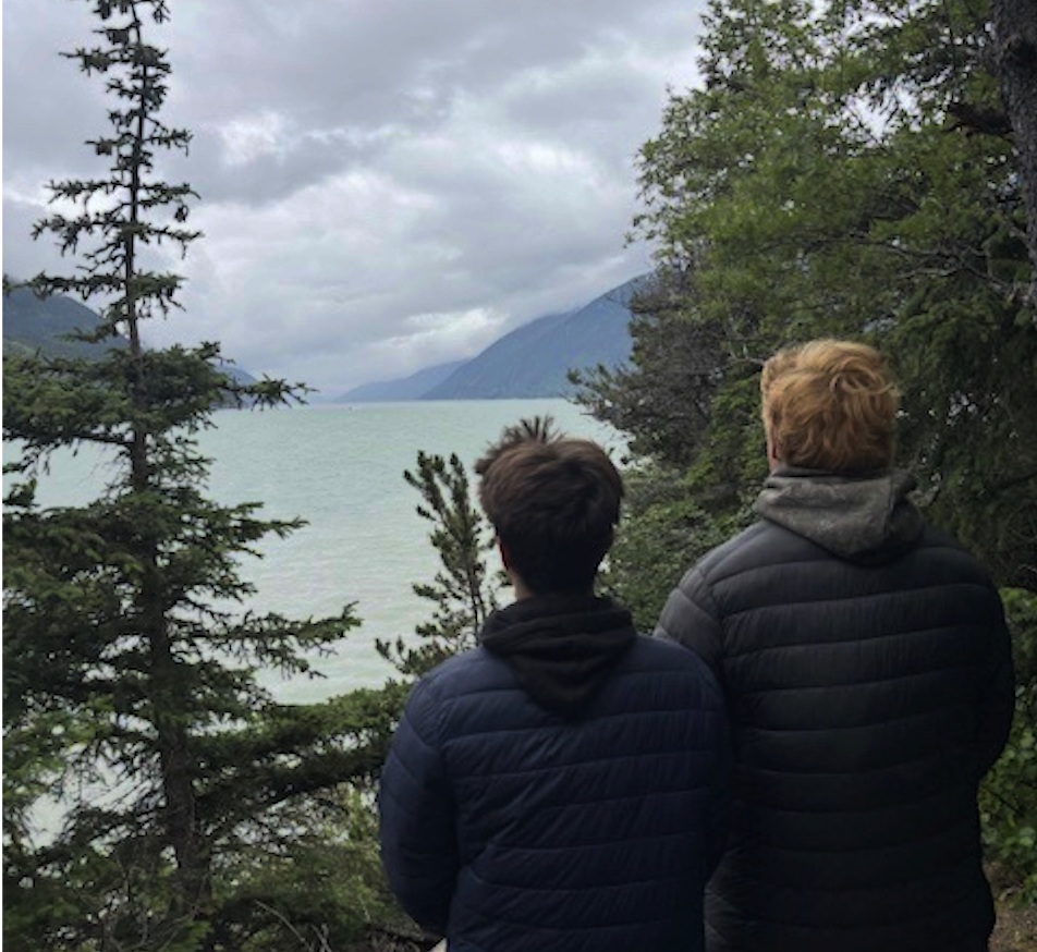 Two people stand on a trail overlooking a large body of water with mountains in the distance and cloudy skies, surrounded by trees and bushes.