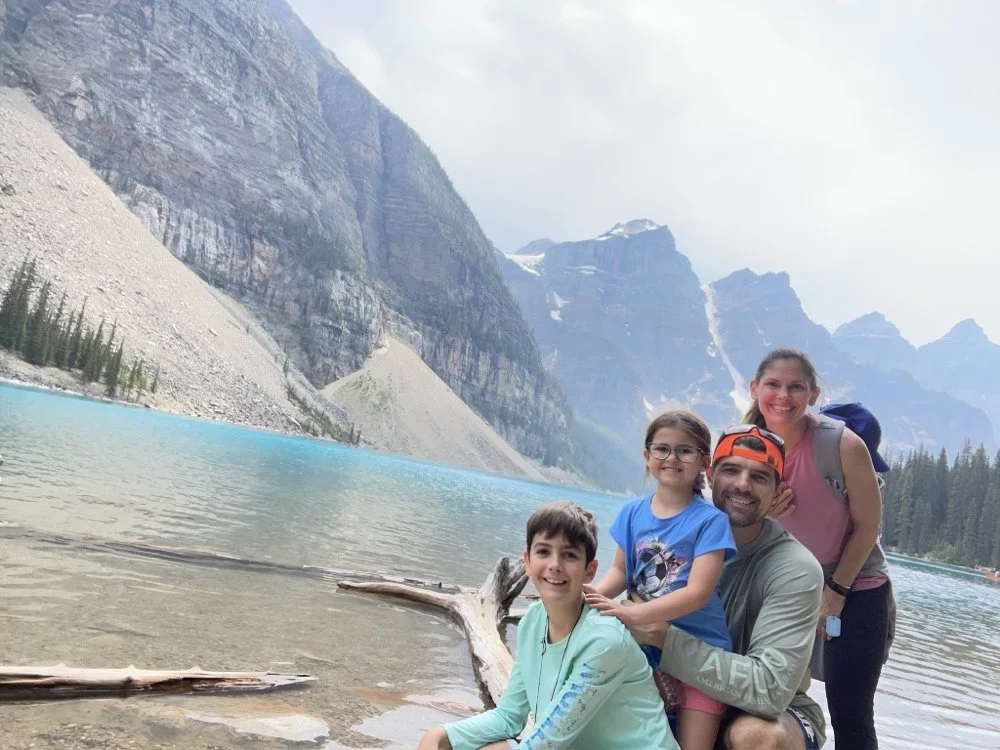 A family of four posing at Moraine Lake in a mountainous national park. The family consists of two children, a girl and a boy, and two adults, a man and a woman. They are smiling with mountains and a bright blue lake in the background.