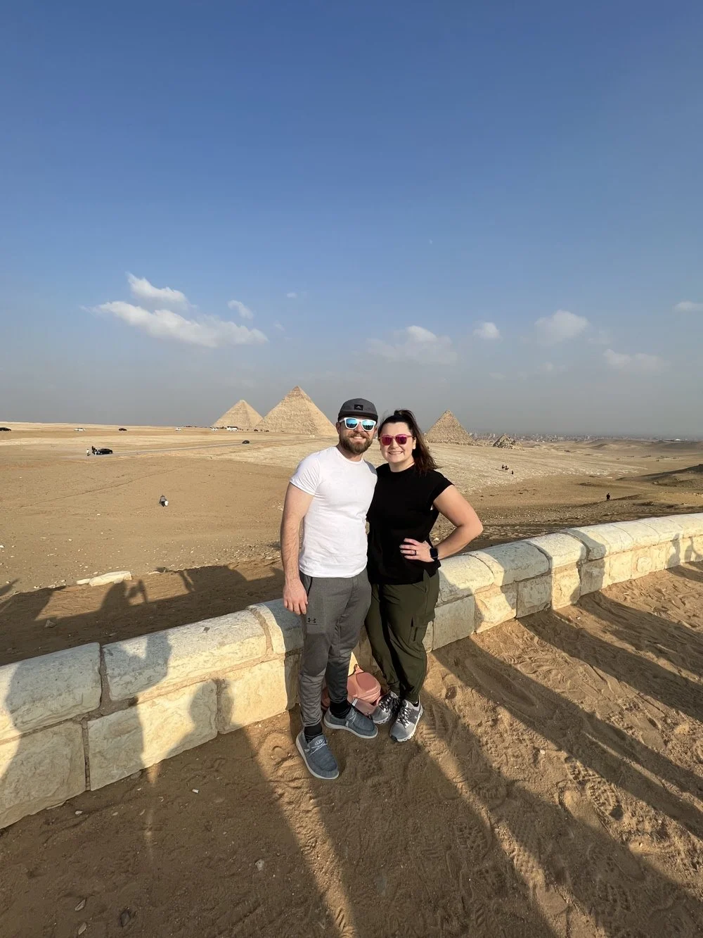 A couple poses for a photo in front of the Pyramids of Giza in Egypt on a sunny day, standing beside a stone barrier with sand around them.
