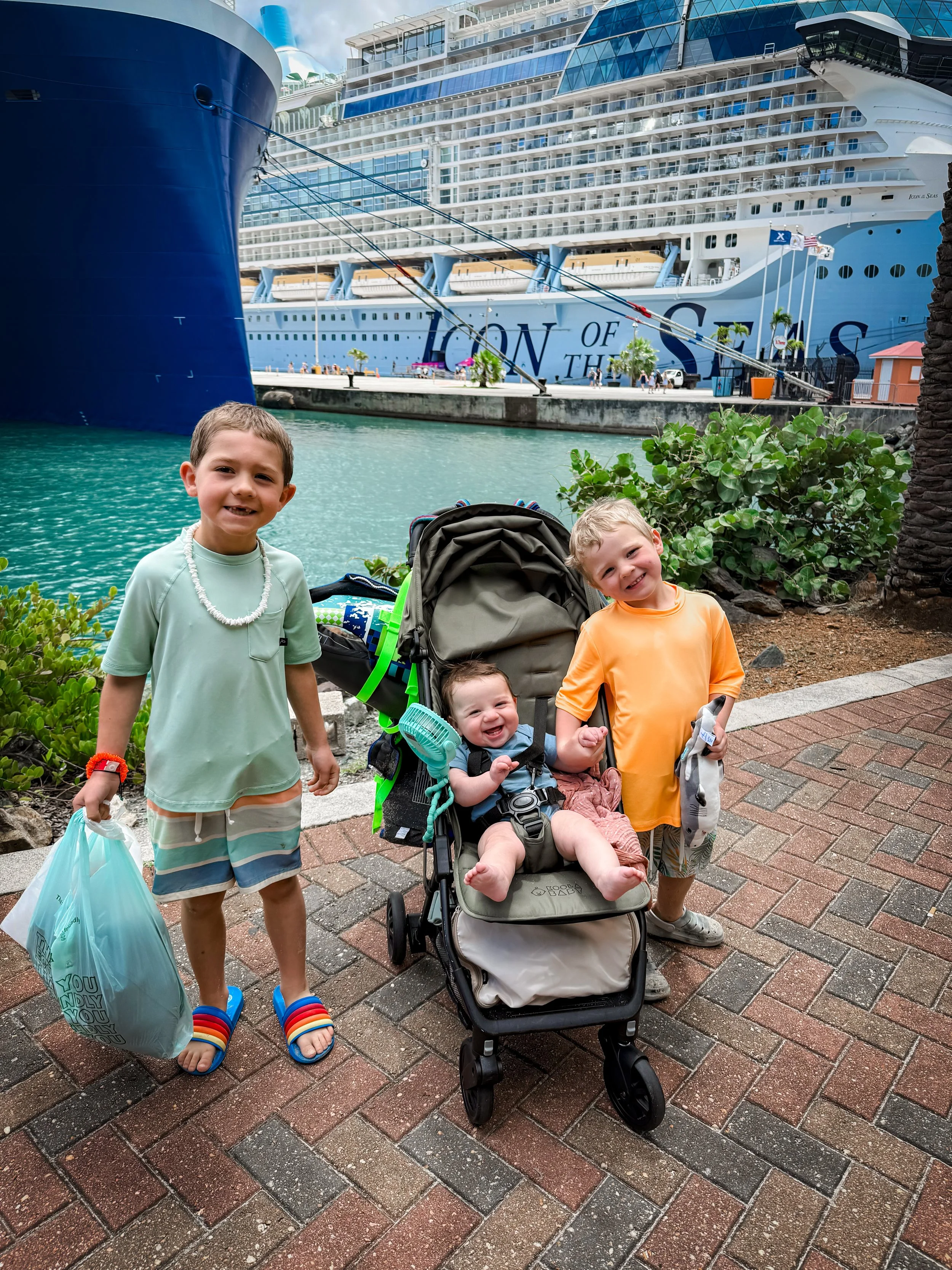 Four children standing on a brick sidewalk in front of a harbor with a large cruise ship named 'Icon of the Seas' docked behind them. Two children are standing, one is sitting in a stroller, and one is partially visible sitting in the stroller. The children are smiling and dressed in casual summer clothes, some holding bags and toys.
