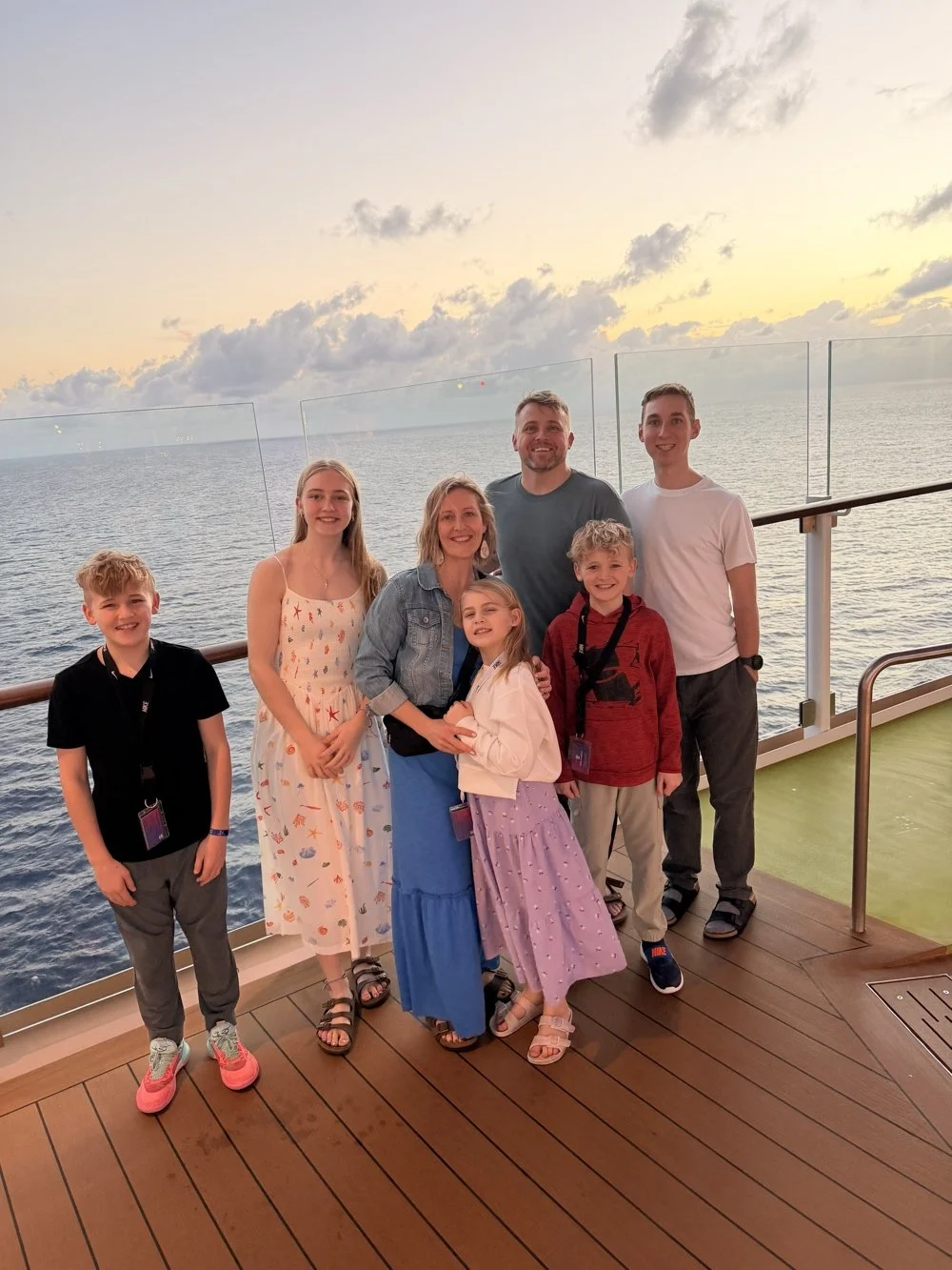 A family of seven stands on the deck of a cruise ship during sunset, smiling at the camera with the ocean and cloudy sky in the background.