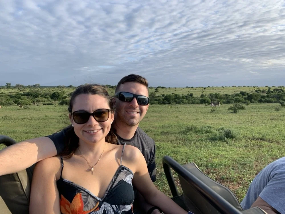 Two people sitting in a safari vehicle, smiling at the camera, with a vast savannah and wildebeest in the background on a cloudy day.