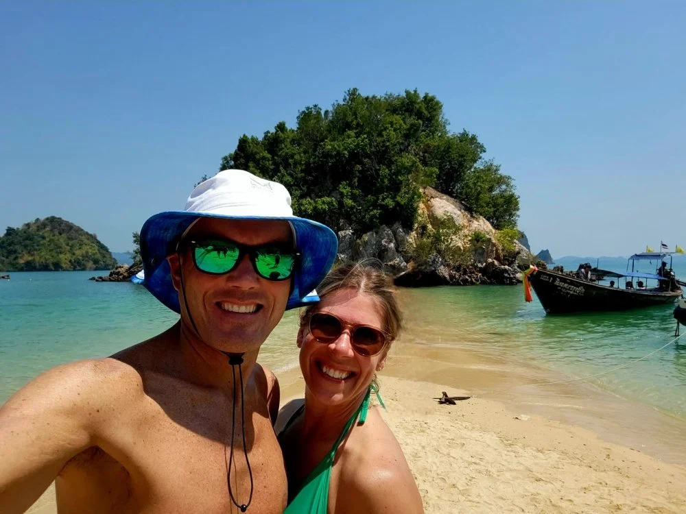 A smiling man and woman taking a selfie on a beach with clear water, a small island with trees, and a boat in the background.