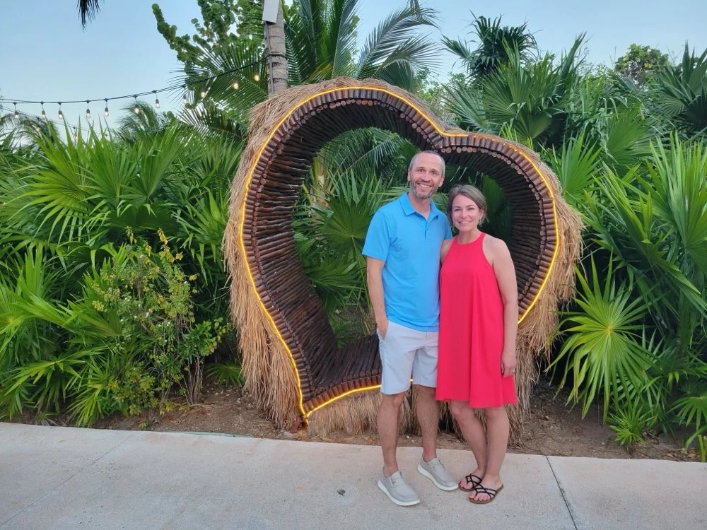 A smiling couple standing outdoors in front of a heart-shaped wooden and straw decoration with tropical plants.