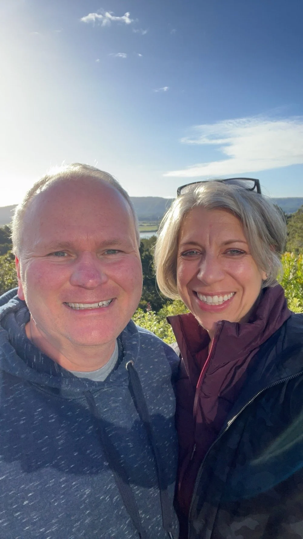 A smiling man and woman taking a selfie outdoors on a sunny day, with green foliage and a blue sky with some clouds in the background.