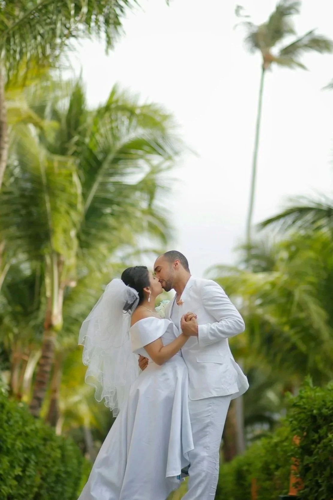 A newlywed couple dressed in white, sharing a kiss and dance outdoors among palm trees.
