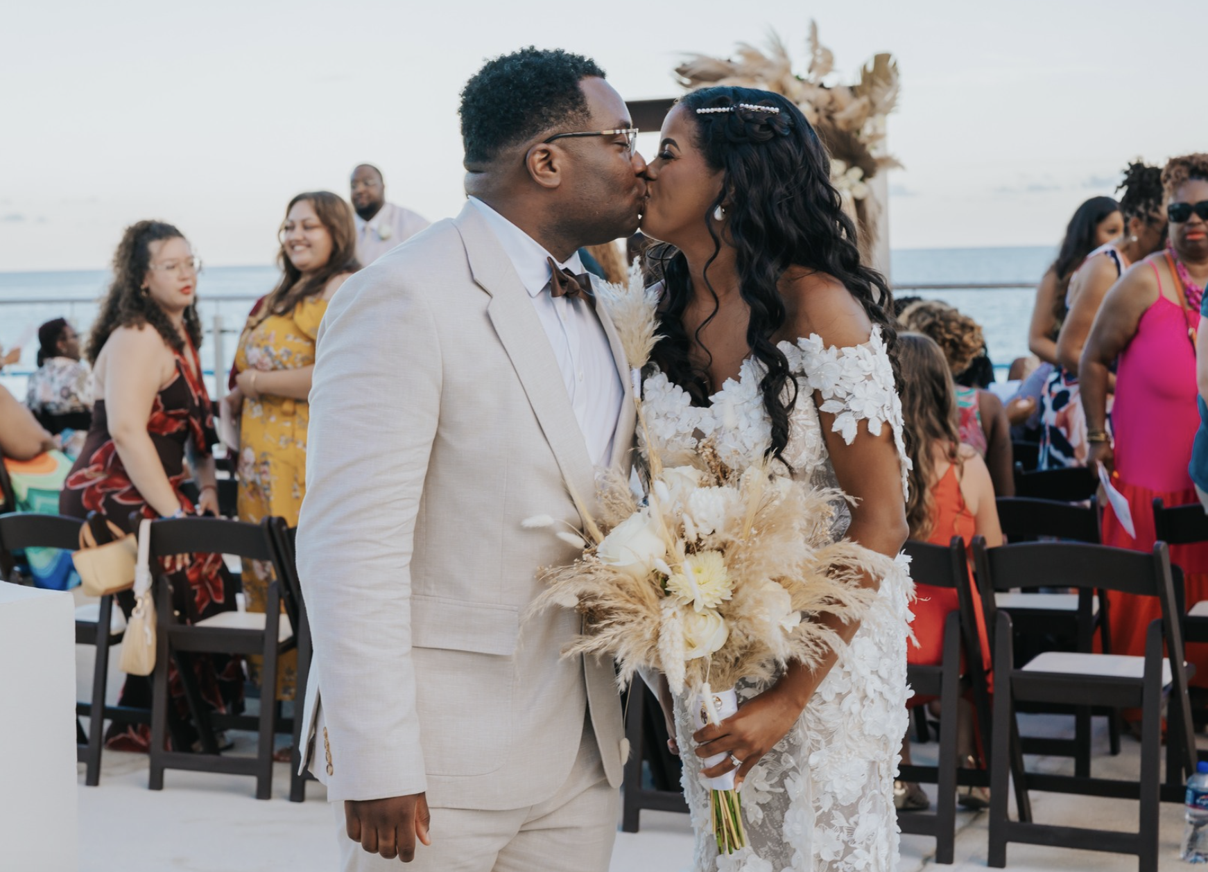 A couple is sharing a kiss at their wedding ceremony on a beach, surrounded by guests and ocean views.