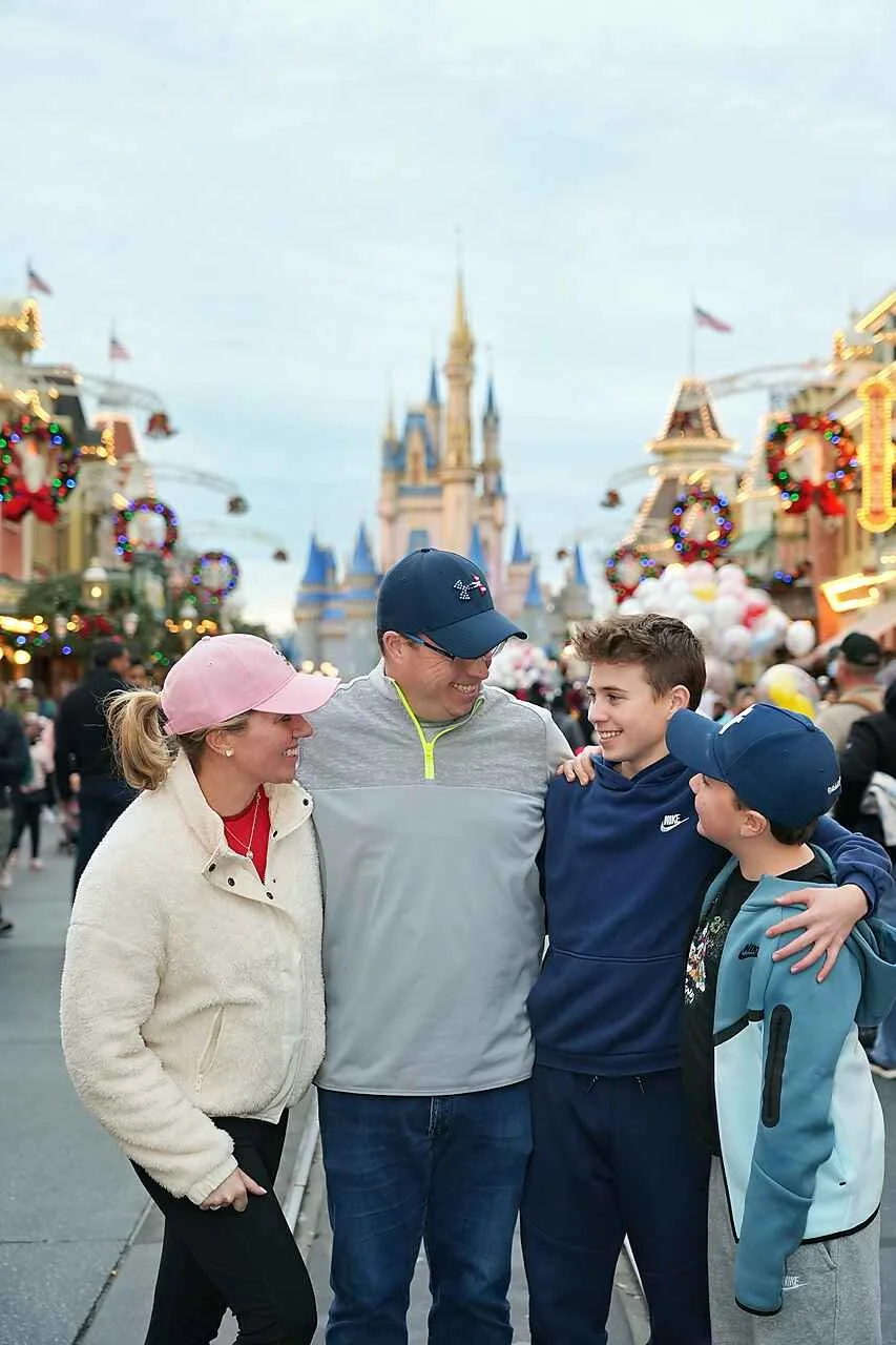A family of four smiling and talking at a Disney theme park, with Cinderella's Castle in the background, decorated for Christmas with lights and wreaths.