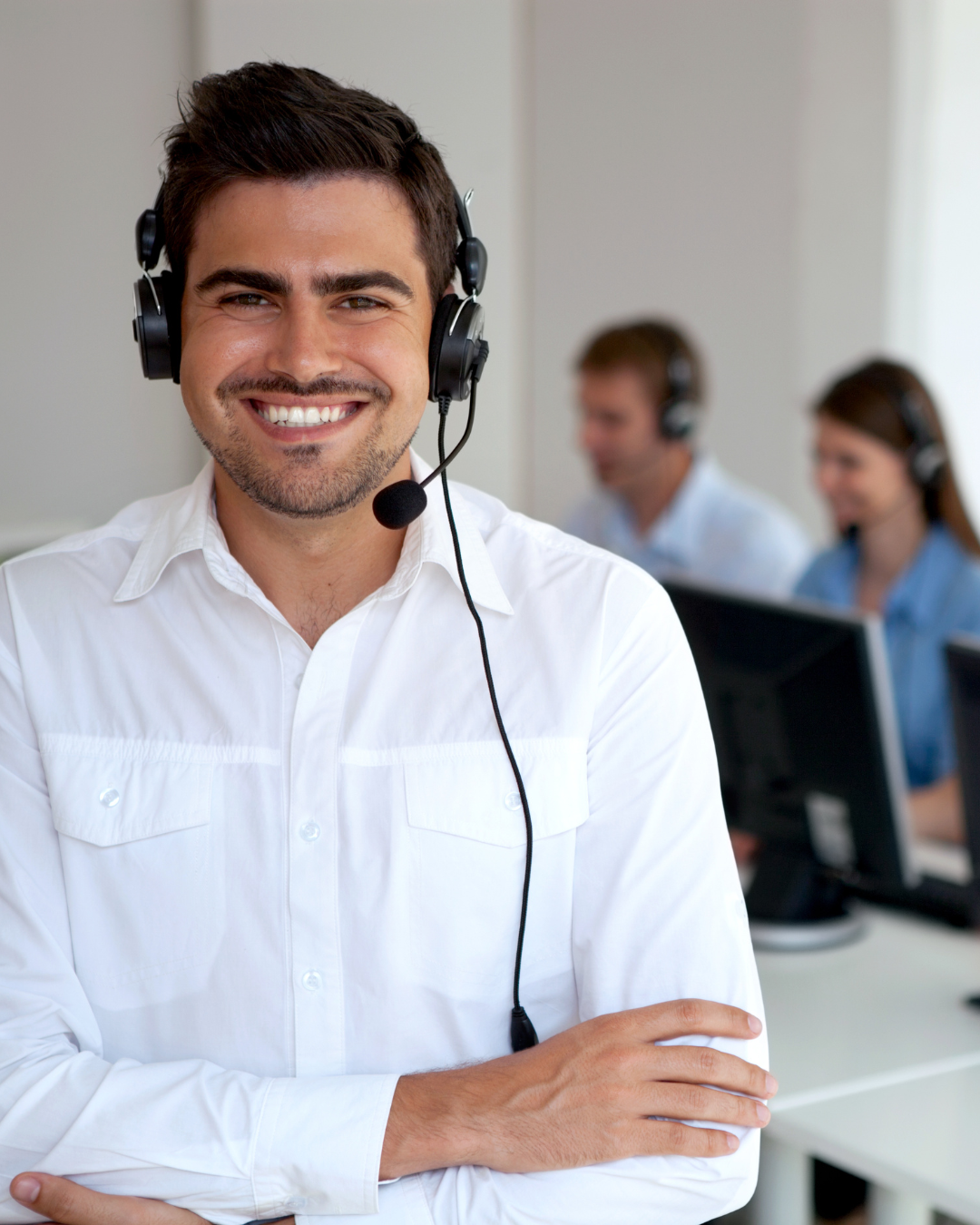 En man med vit skjorta som ler och bär headset i ett callcenter, med två andra personer i bakgrunden som också jobbar vid datorer och har headset.
