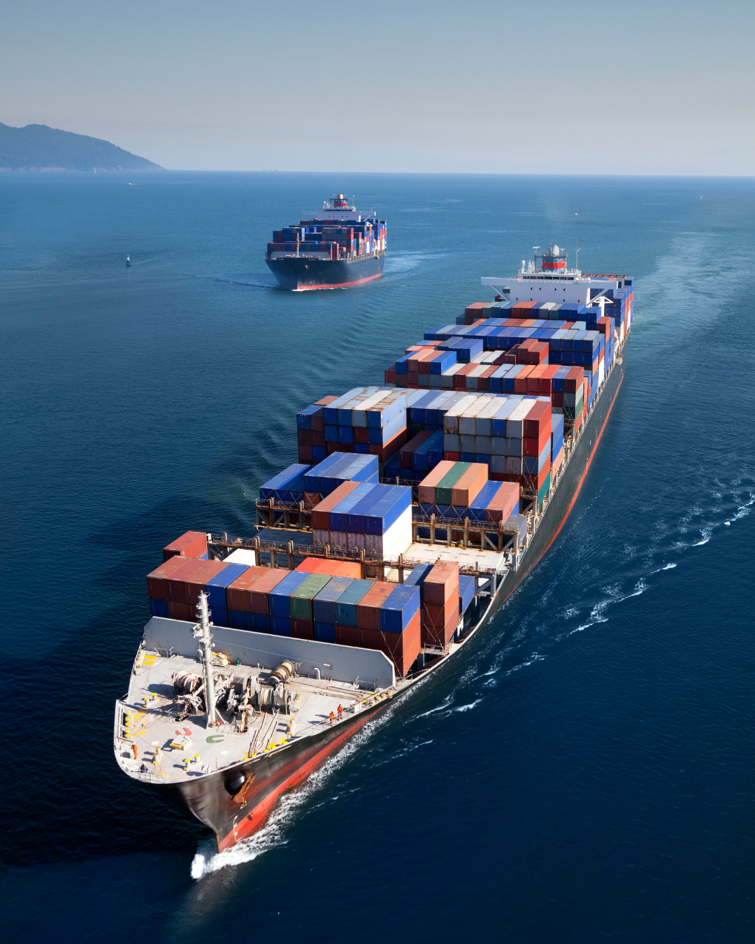 Två stora containerfartyg som seglar på havet, fyllda med färgglada containrar, med en landmassa i bakgrunden.
