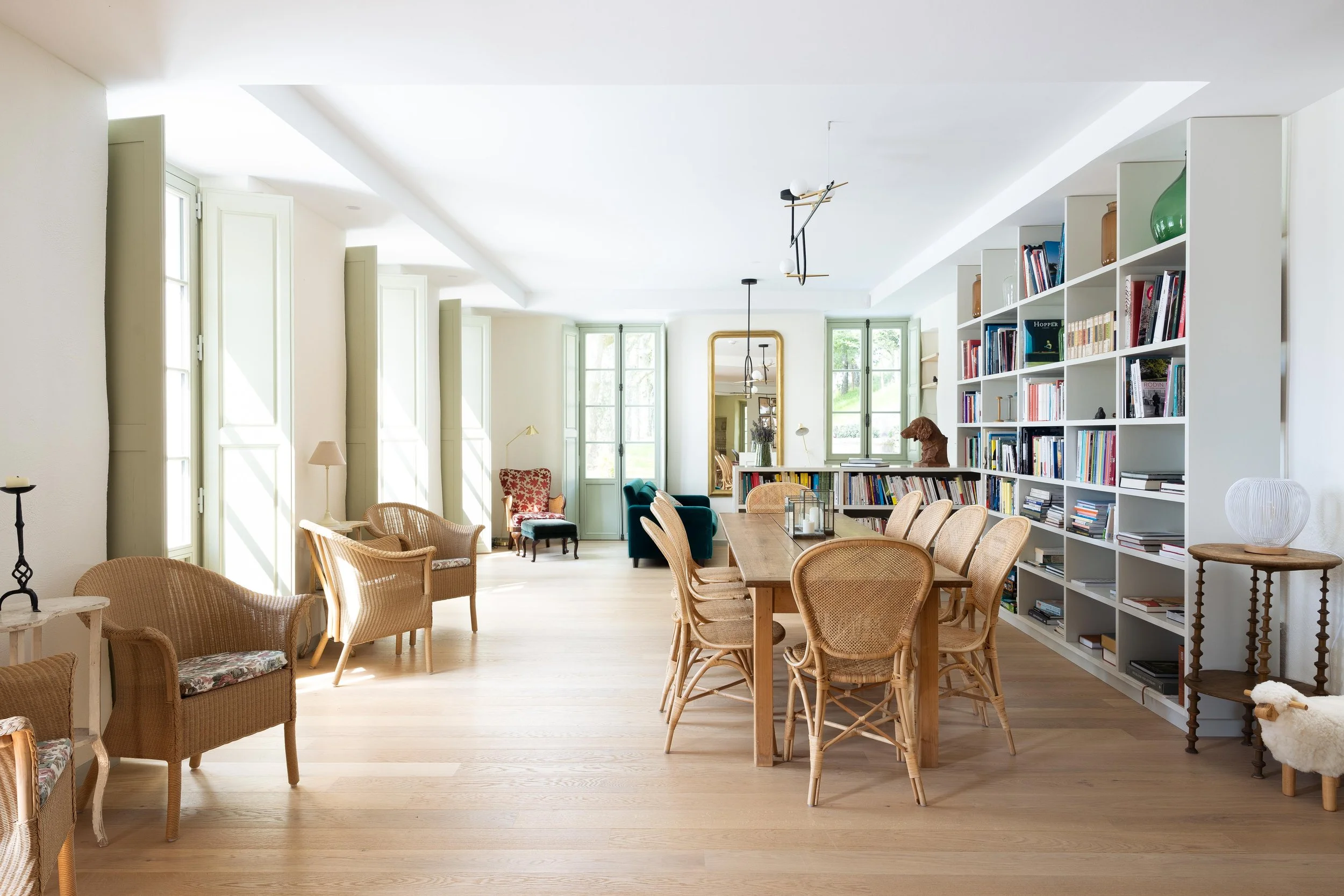 Salle de lecture lumineuse avec un grand plafond blanc, des fenêtres ouvertes, des étagères remplies de livres, des fauteuils en osier et des tables en bois.