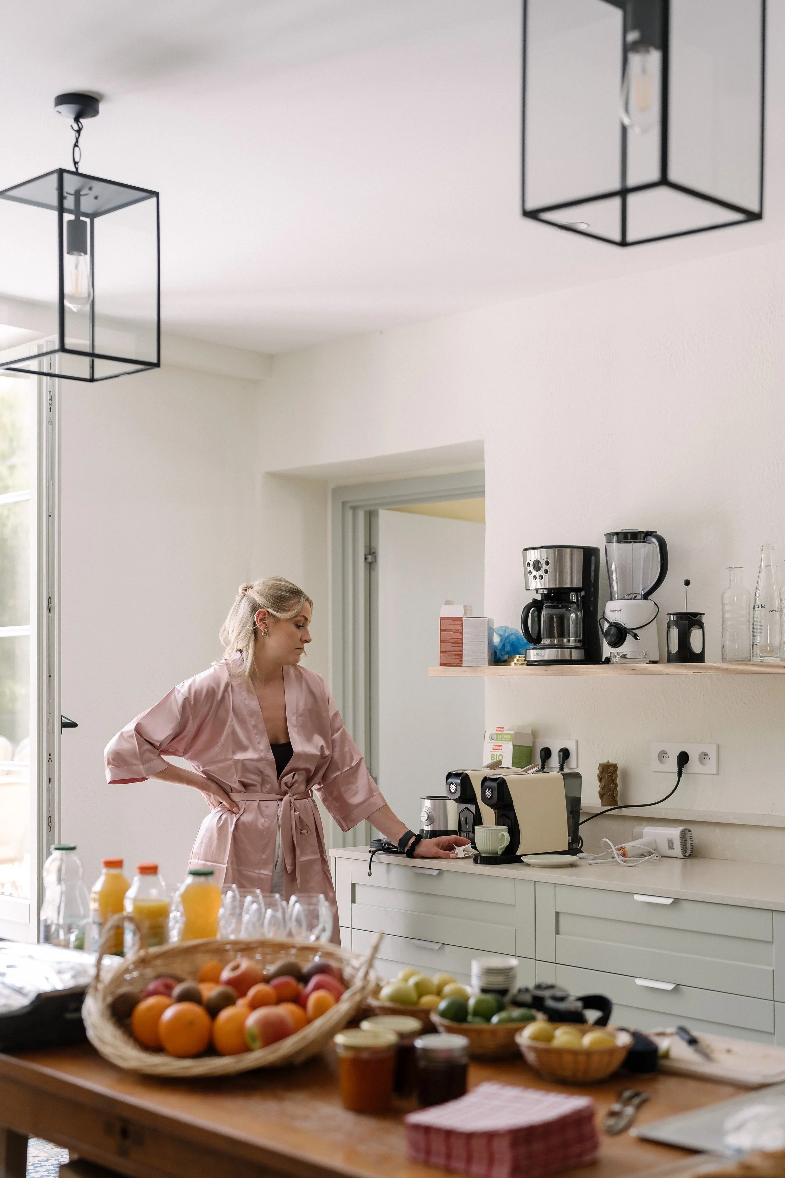 Une femme debout dans une cuisine blanche près d'une cafetière, portant une robe rose avec une ceinture, entourée de paniers de fruits et de petits pots de confiture.