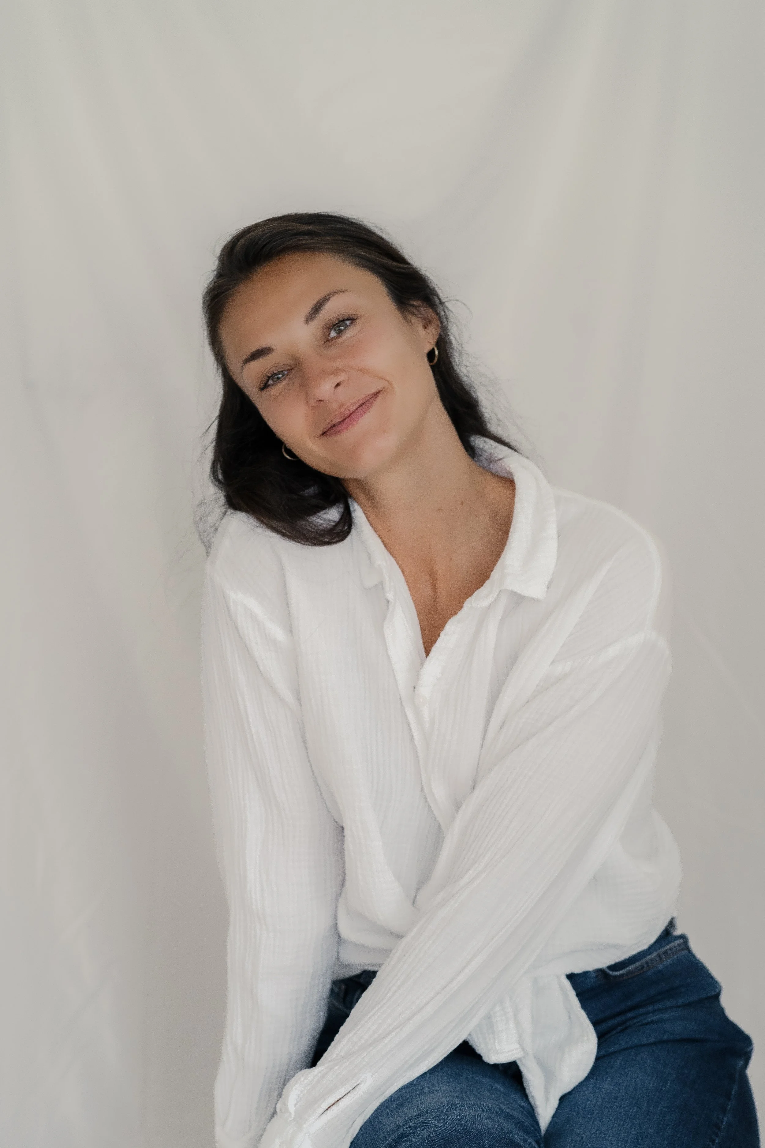 Ein junger Frau mit braunen Haaren, die lächelt und neben einer weißen Wand sitzt, trägt eine weiße Bluse und blaue Jeans.