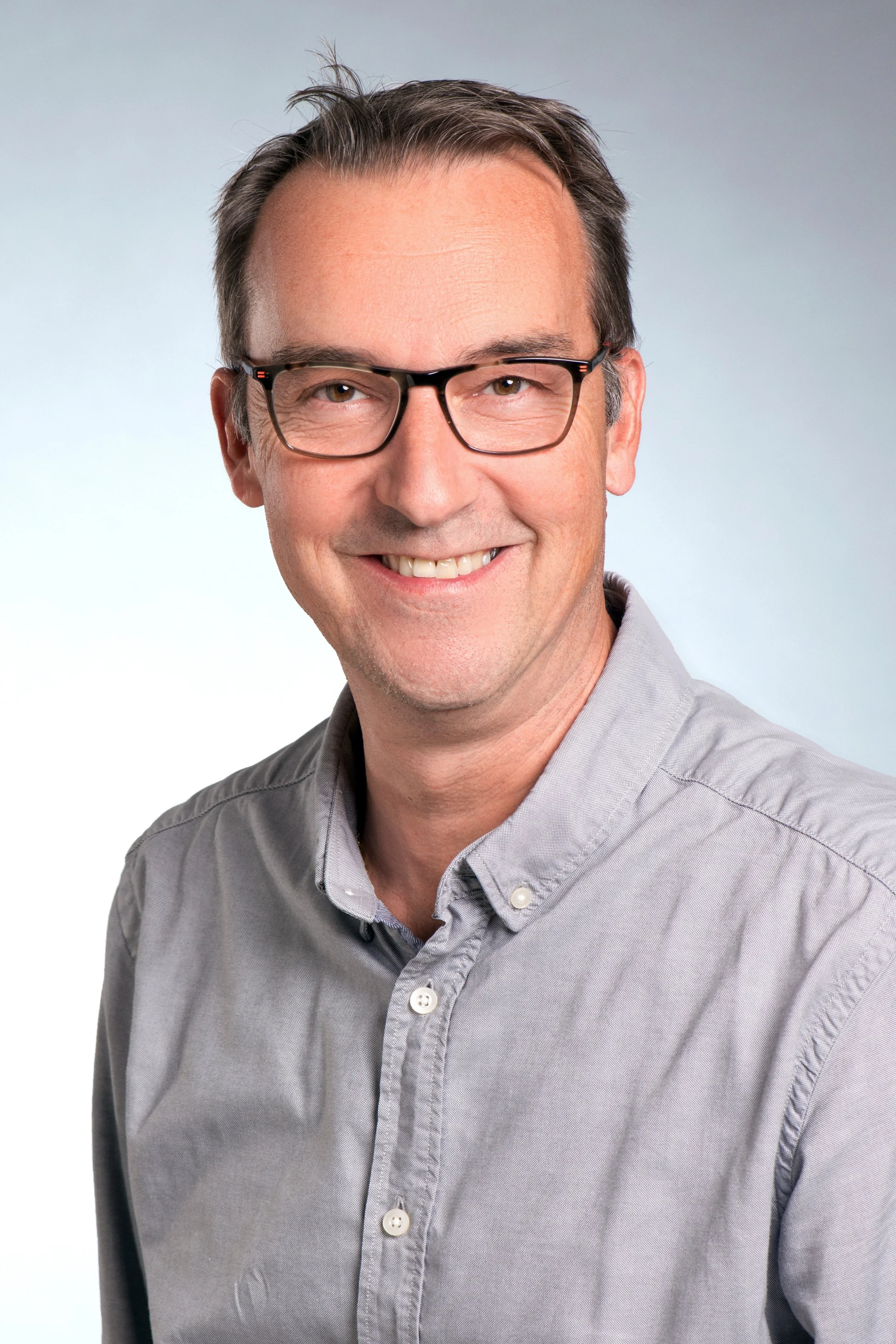 Ein lachender Mann trägt eine graue Hemd und schwarze Brille, die Haare sind grau und nach hinten gestylt, der Hintergrund ist hell.