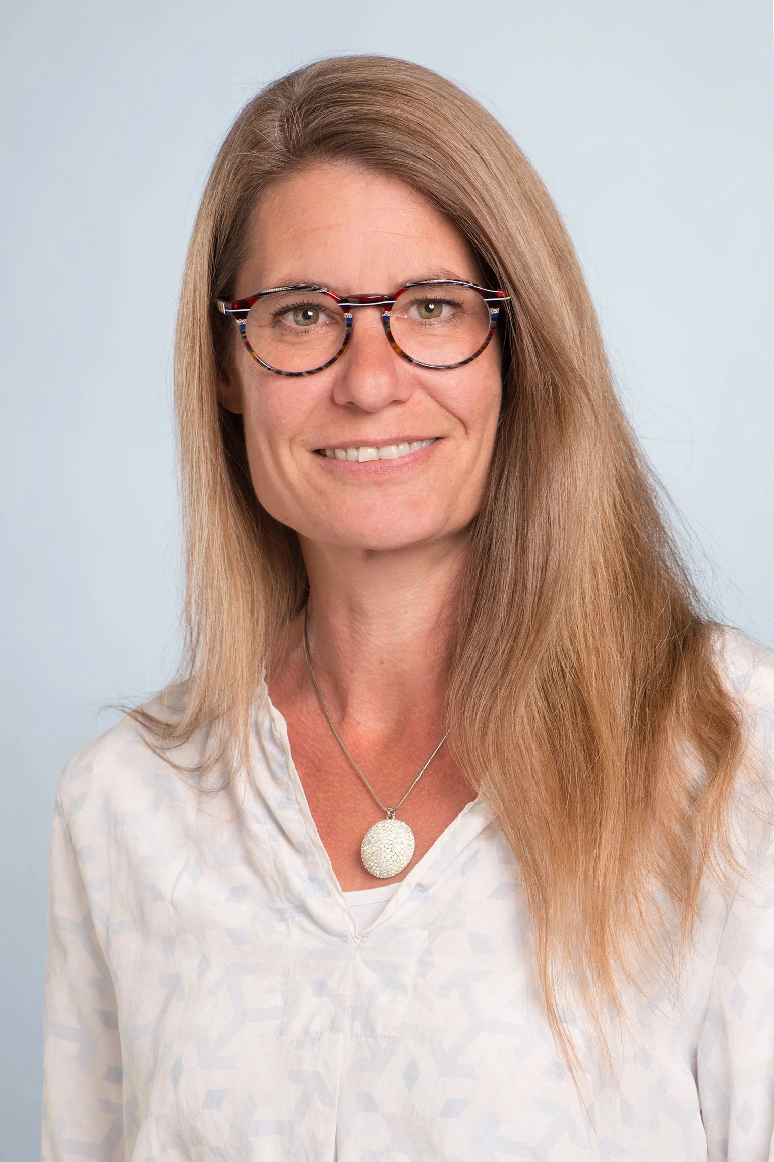 Eine Frau mit langen, hellbraunen Haaren trägt schwarze Brillen mit bunten Mustern, ein weißes Hemd und eine silberne Halskette mit einem großen, runden Anhänger.