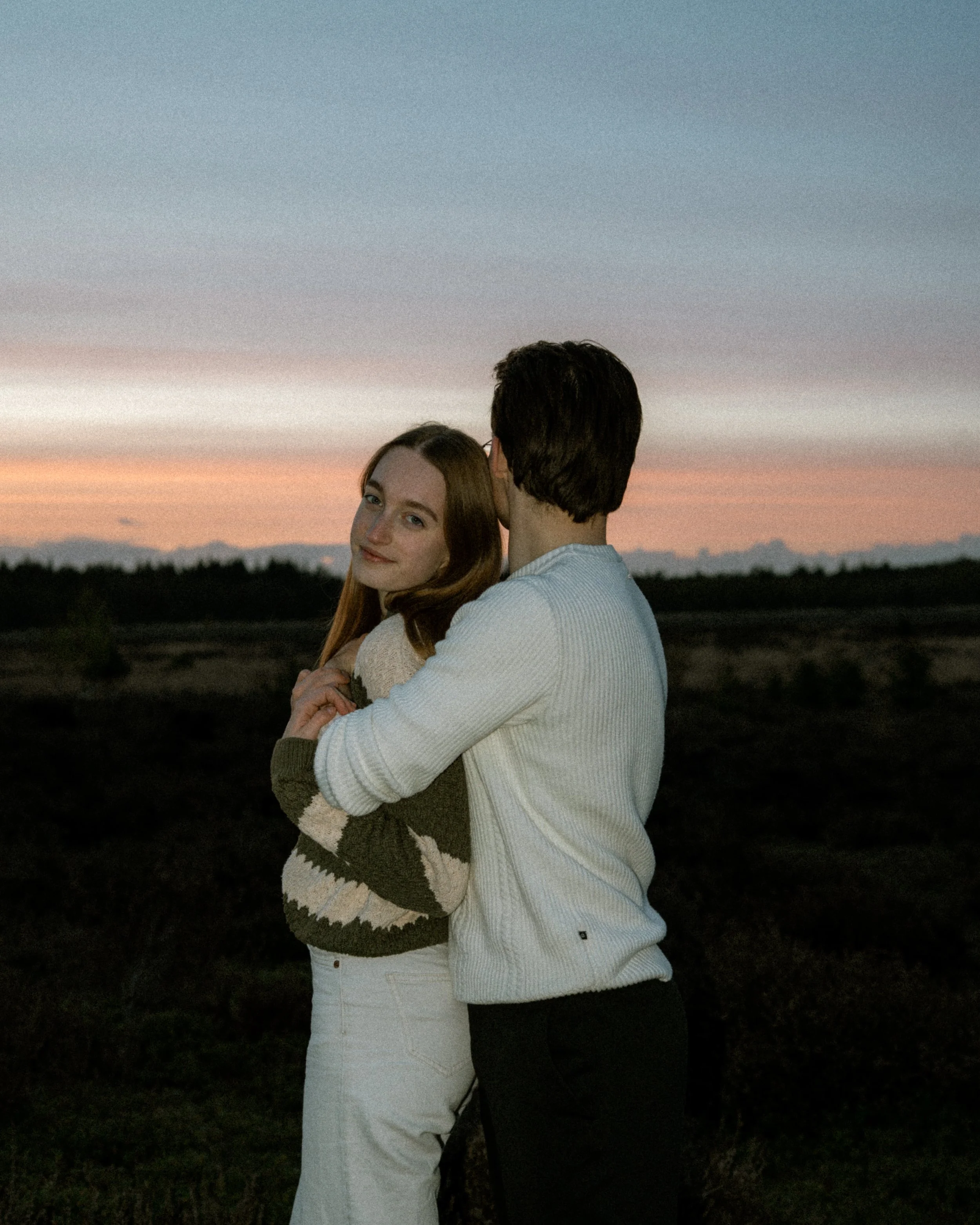 Een portret van een jong stel, arm in arm, staan in een veld tijdens zonsondergang. De vrouw kijkt blij en naar de camera, de man kijkt weg, hun gezichten zwart door de silhouet.