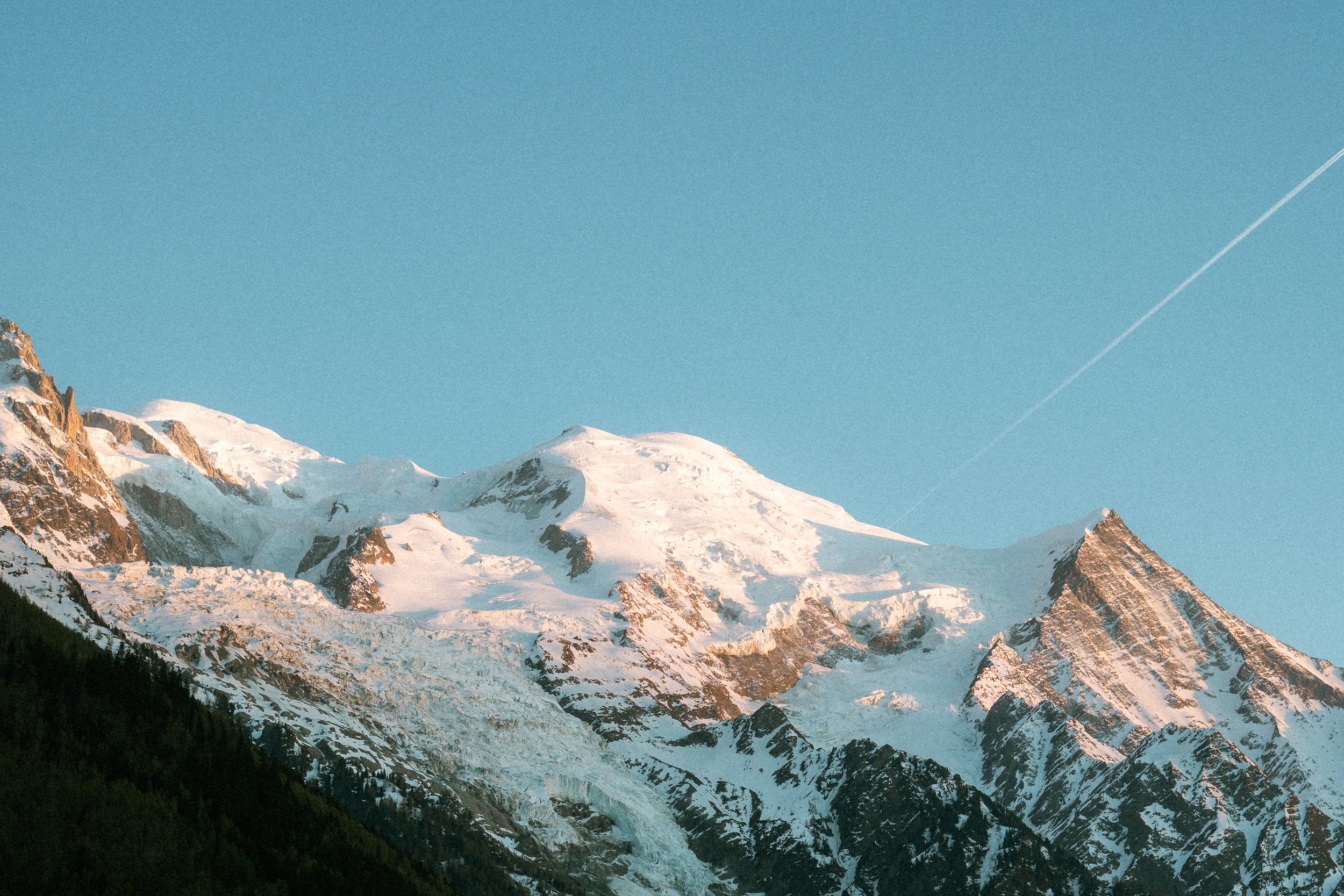 Foto van de sneeuwbedekte bergtoppen van Mont Blanc onder een blauwe hemel met een vliegtuigstrepen in de lucht.