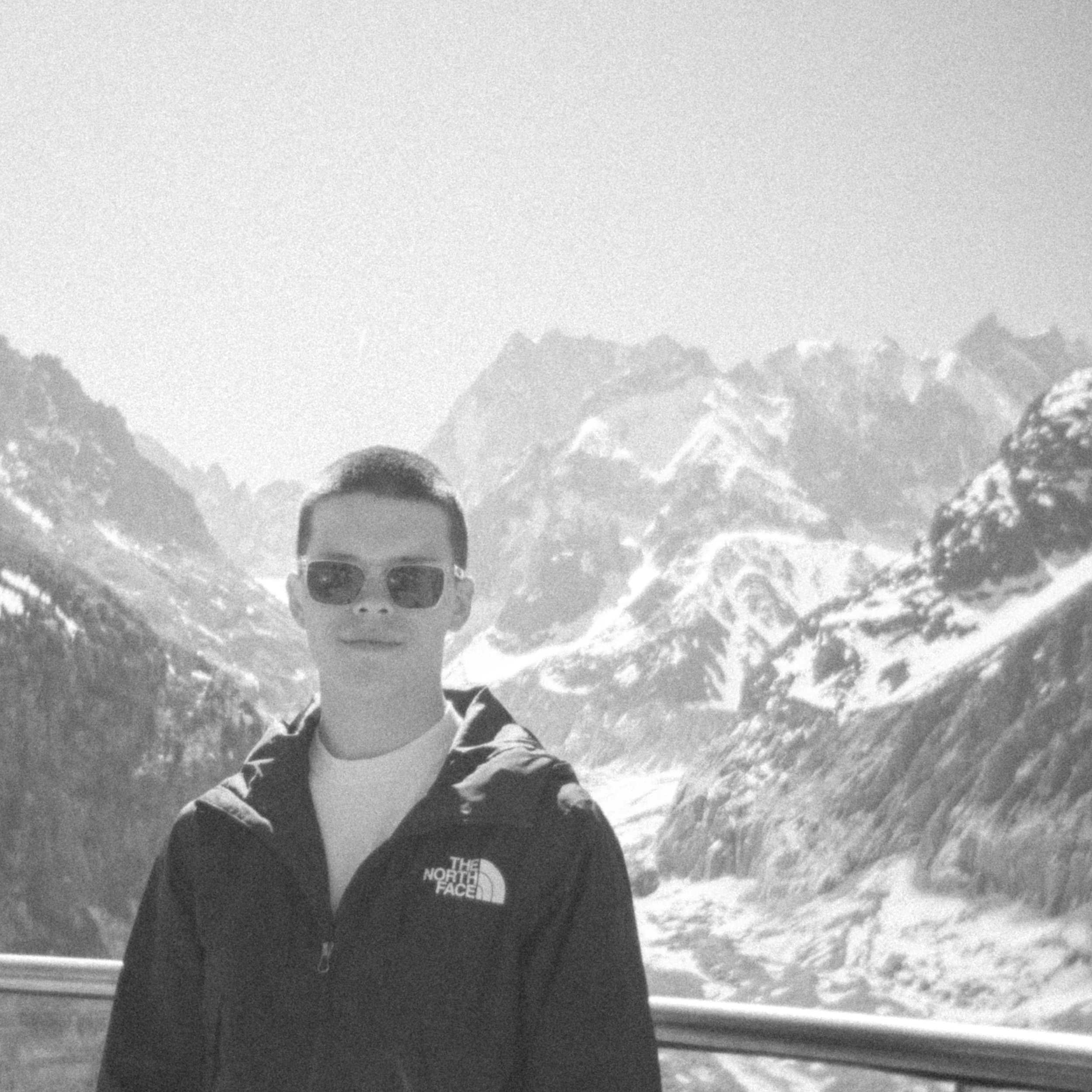 Een persoon met een zwart North Face jasje en een zonnebril staat in een bergachtig landschap, mogelijk in de sneeuw, met besneeuwde bergen op de achtergrond.