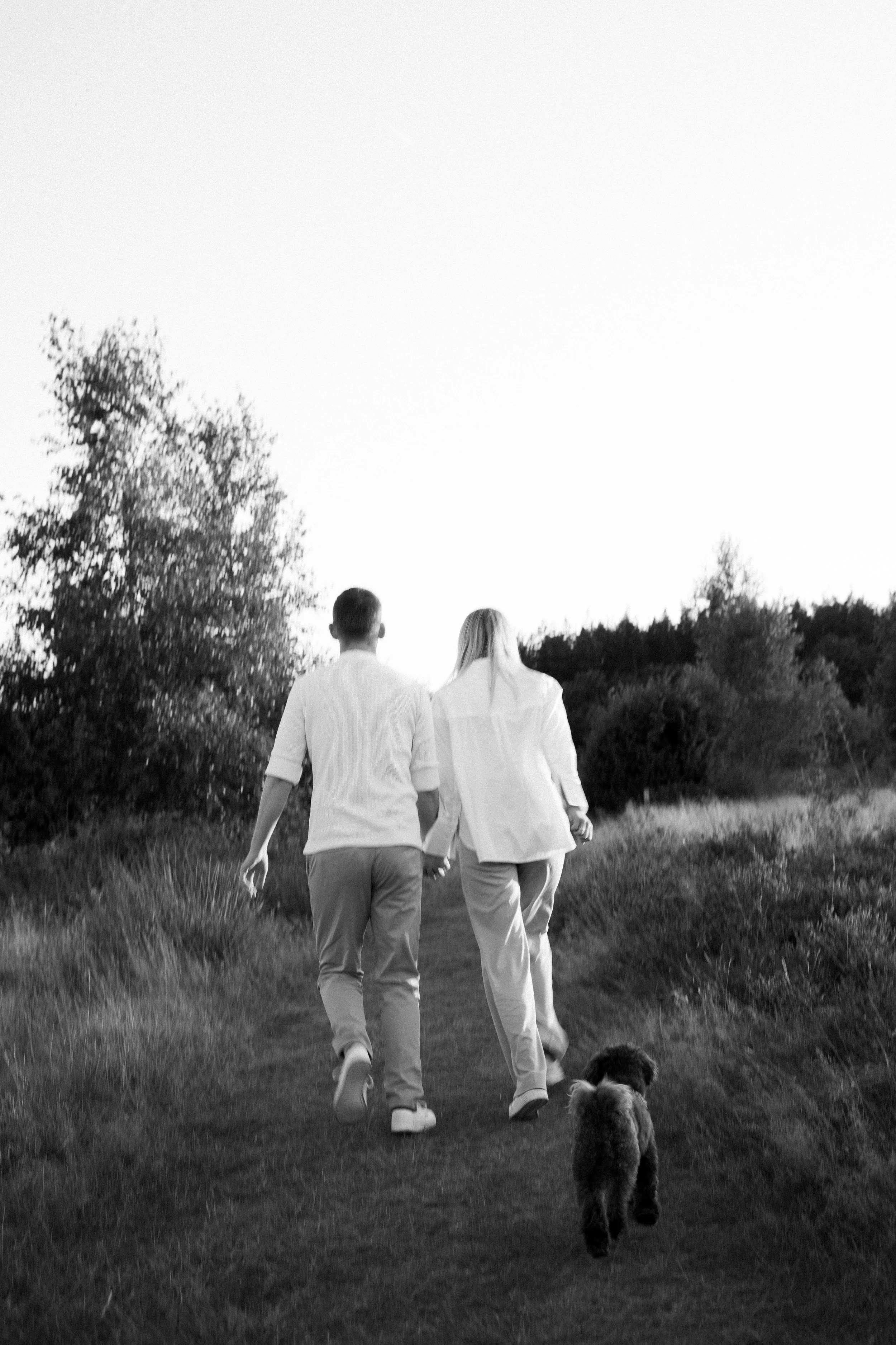 Twee mensen, een man en een vrouw, wandelen hand in hand door een grasveld met bomen op de achtergrond in een zwart-wit foto.