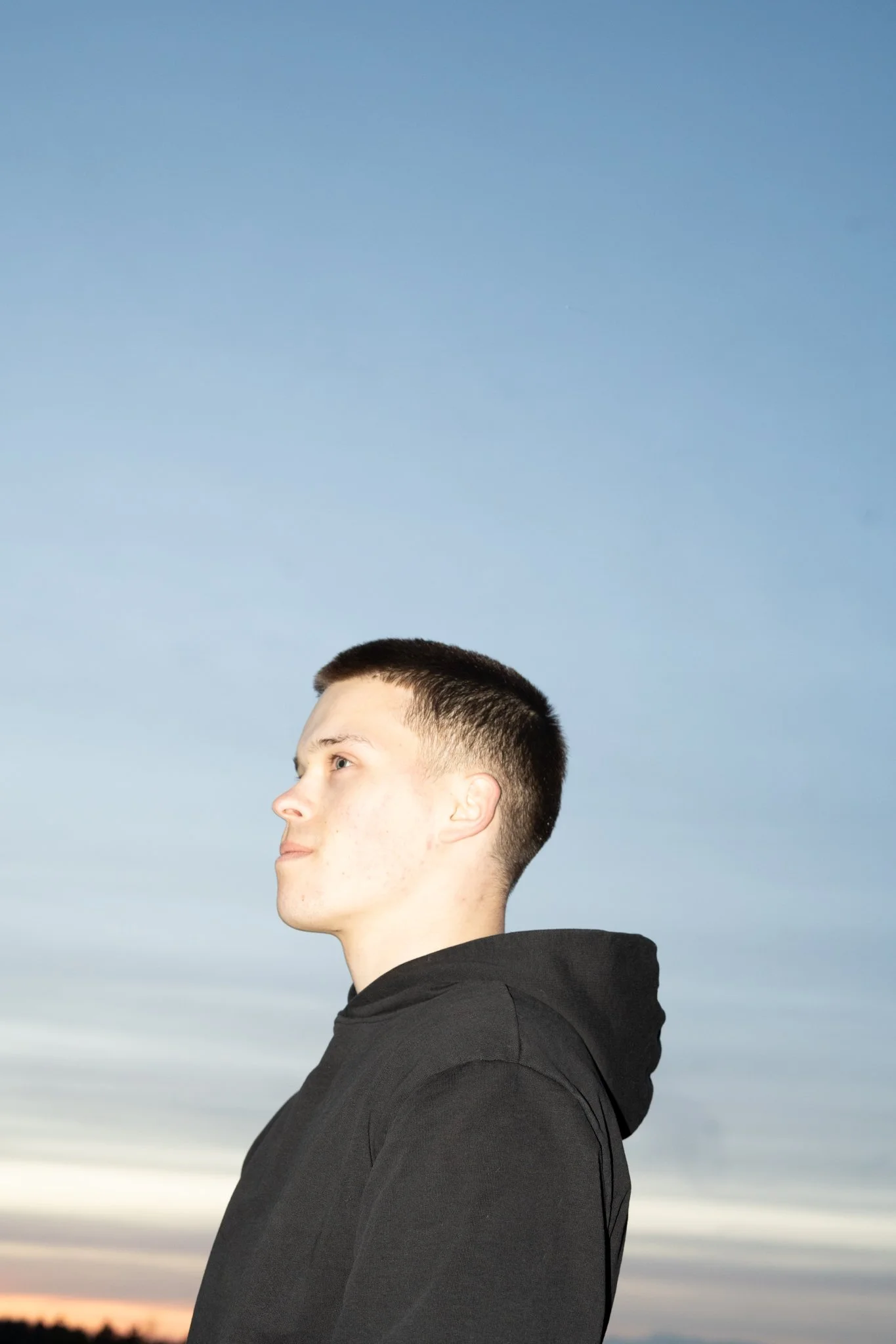 Portret van een jonge fotograaf met kort, donker haar, gekleed in een zwarte hoodie, staande voor een heldere blauwe hemel tijdens zonsopgang of zonsondergang.