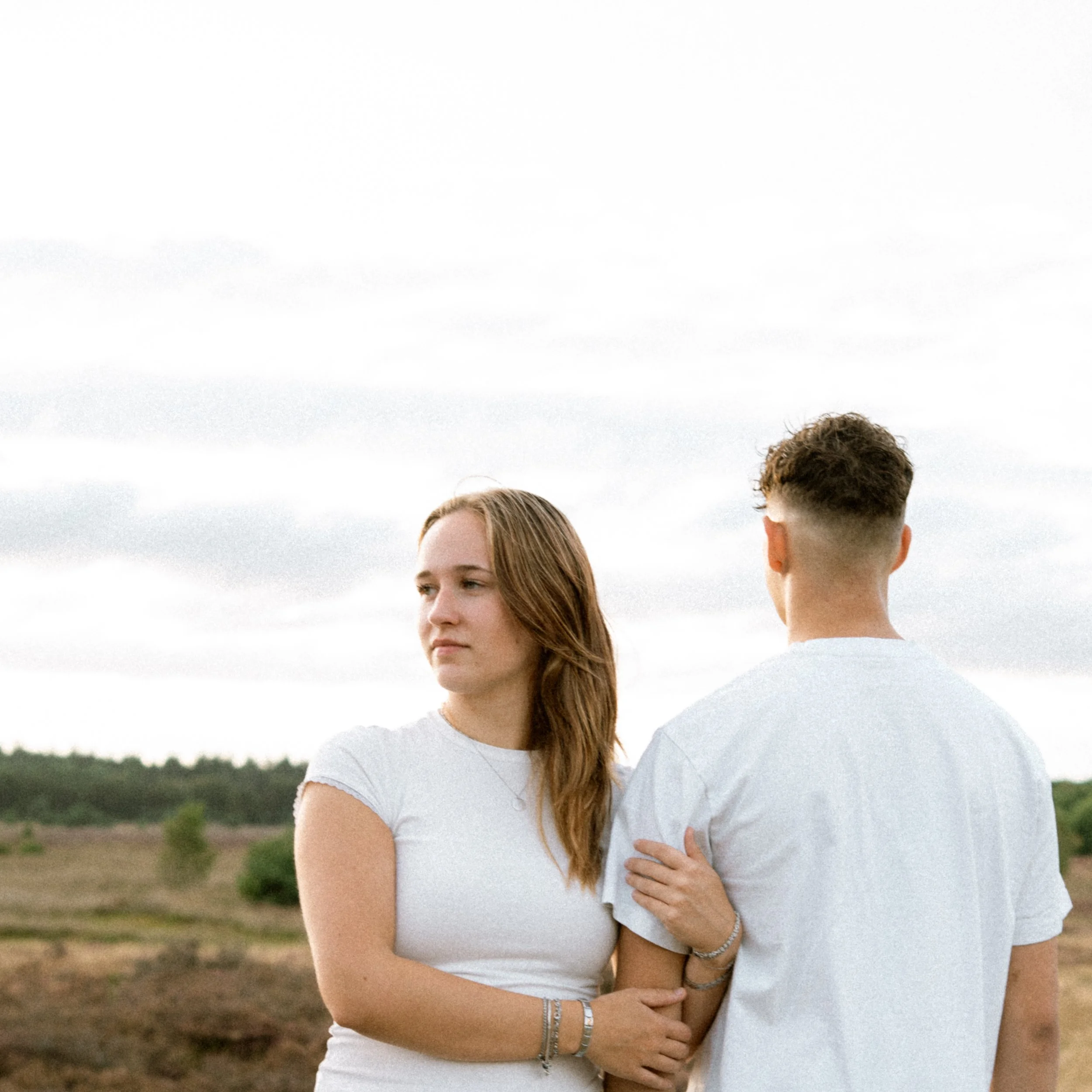Twee mensen, een man en een vrouw, staan dicht bij elkaar in een open veld met een bewolkte hemel op de achtergrond. Ze dragen witte t-shirts en hebben sieraden om hun armen.