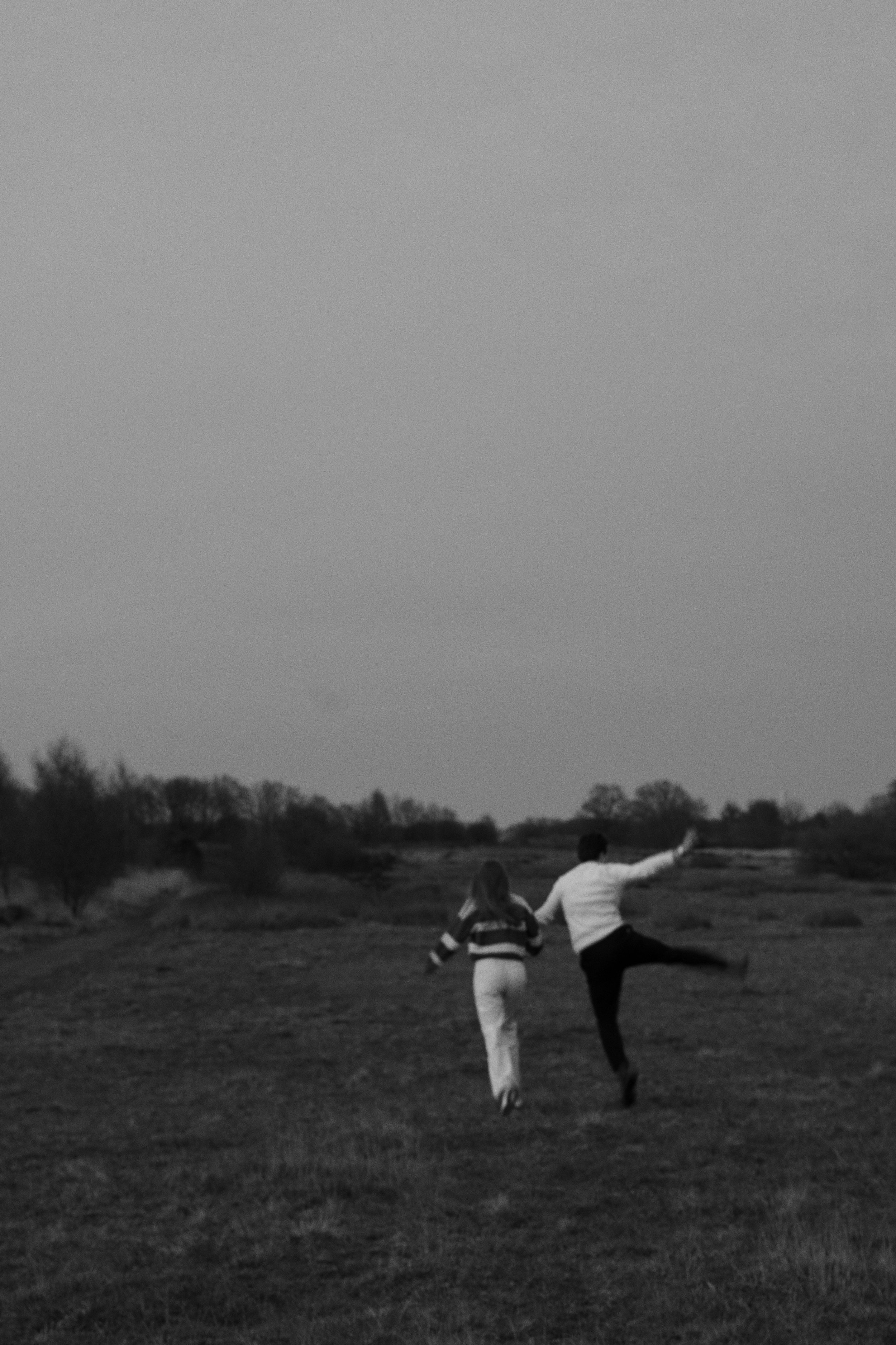Twee mensen dansen en springen op een open veld bij zonsondergang, het landschap is landelijk met bomen op de horizon.