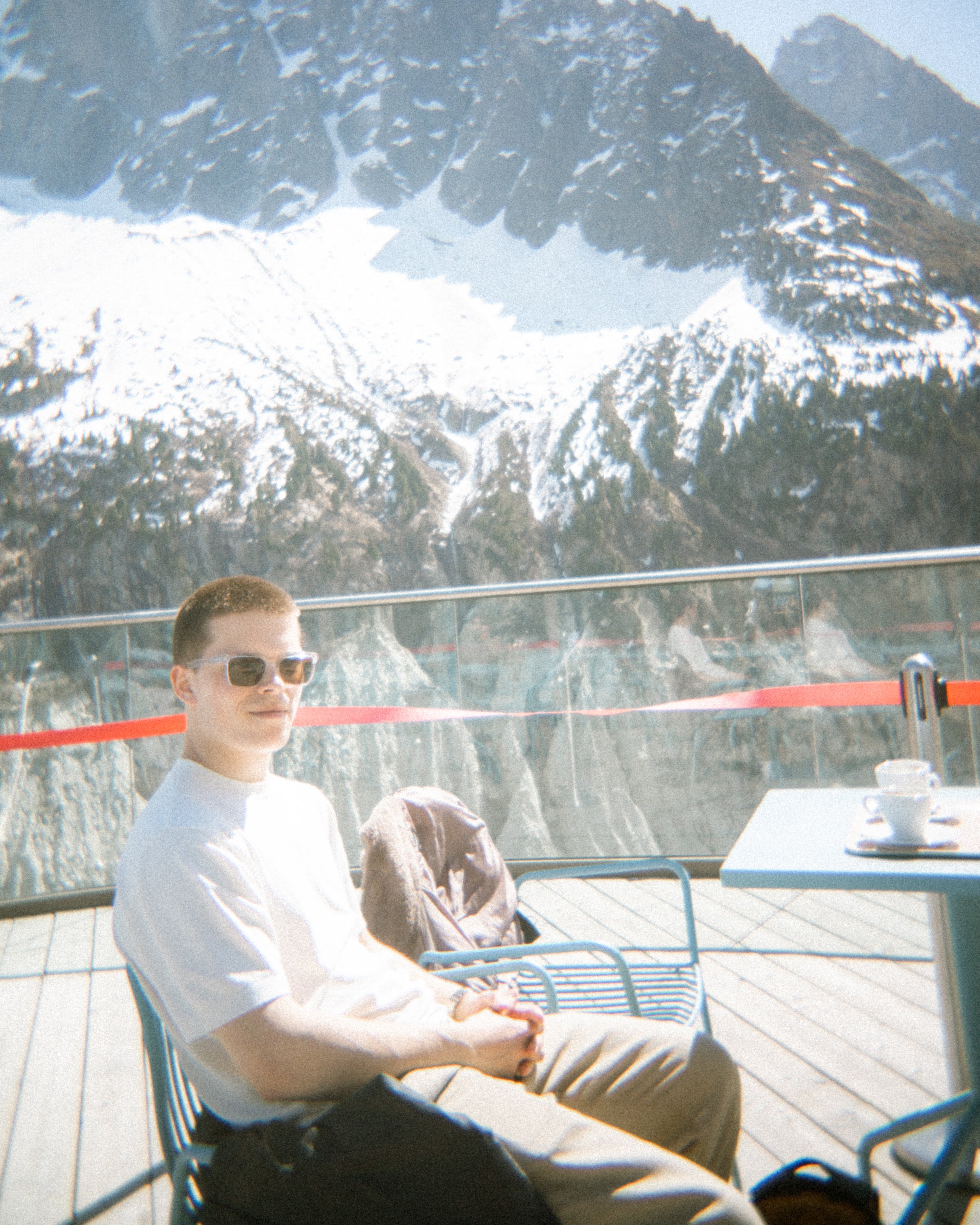 Een jonge man met bruin haar, zwartgekleurde zonnebril, een wit T-shirt en beige broek zit op een blauwe metalen stoel buiten op een terras met uitzicht op bergen met sneeuw. Er staat een tafel met een witte tafelkleed en kopjes op.