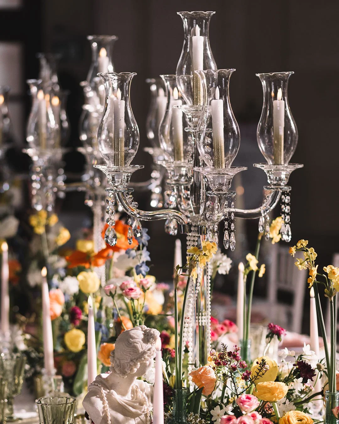Elegant centerpiece featuring a crystal chandelier with candles, surrounded by colorful flowers and a small white bust sculpture.