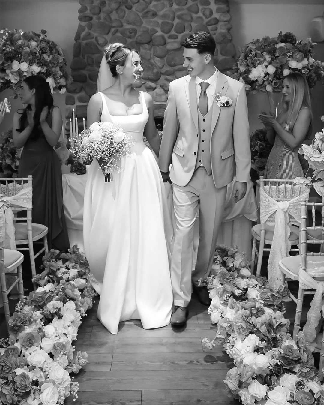 A black and white photo of a bride and groom holding hands and smiling at each other during their wedding ceremony, surrounded by flowers and guests.