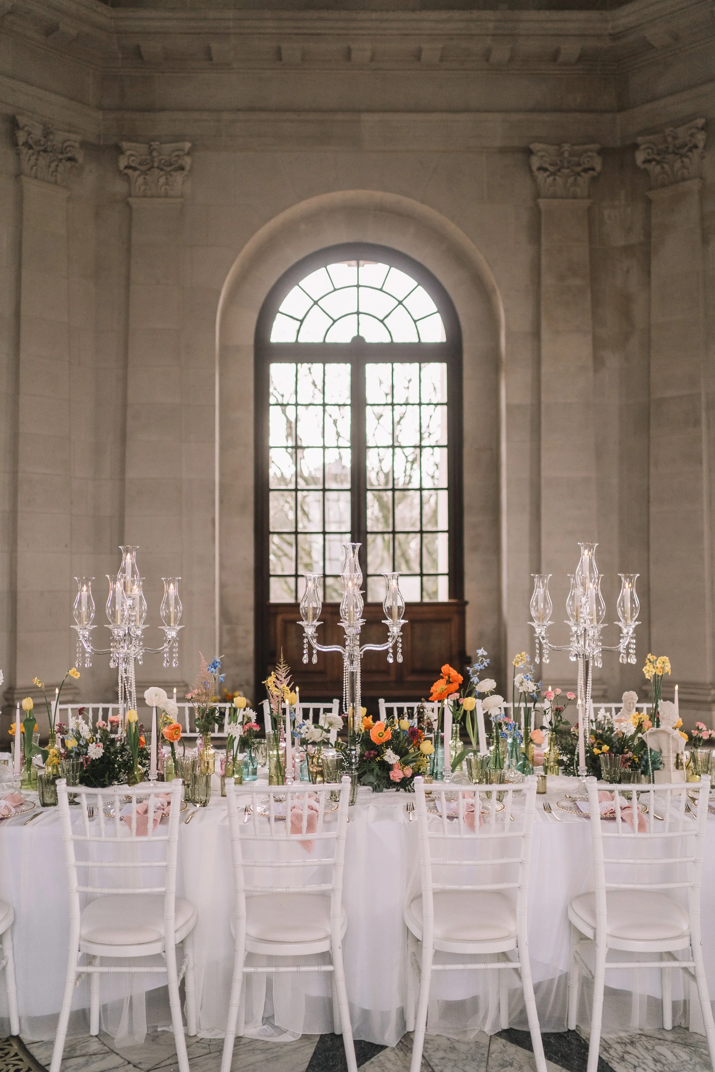 Elegant banquet table decorated with colorful flowers and tall crystal candelabras, set in a grand room with large arched window and classical architectural details.