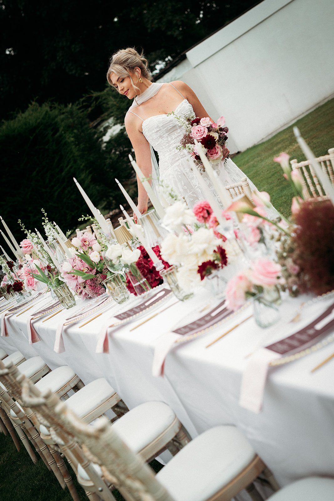 A bride in a white wedding dress standing at a decorated table outdoors, with floral centerpieces and candles, on green grass next to a white wall.