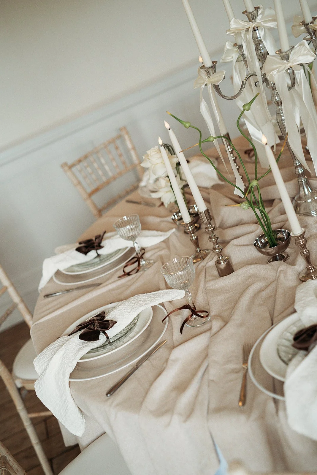 Elegant dining table decorated with white candles, white flowers, glassware, and beige tablecloth for a formal event.
