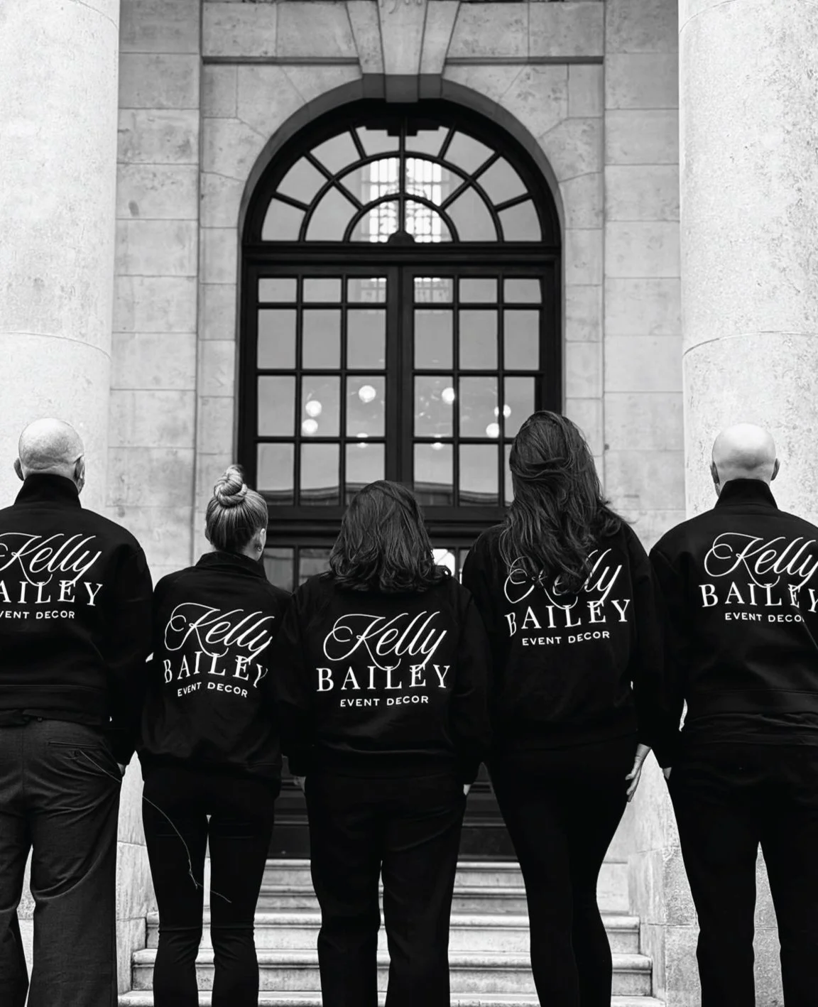 Five people standing on stairs in front of a large window, facing away from the camera. They are wearing matching jackets with "Kelly Bailey Event Decor" written on the back.