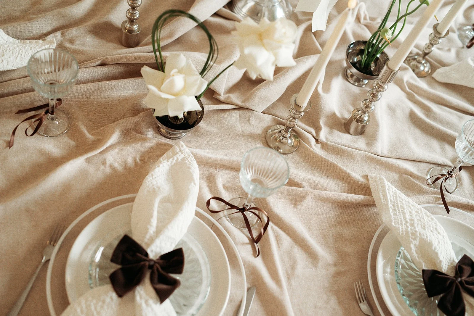 Elegant table setting with beige tablecloth, white plates, cloth napkins tied with dark brown ribbons, clear glassware with ribbons, and tall silver candlesticks with white candles, accented with small black vases of white flowers.