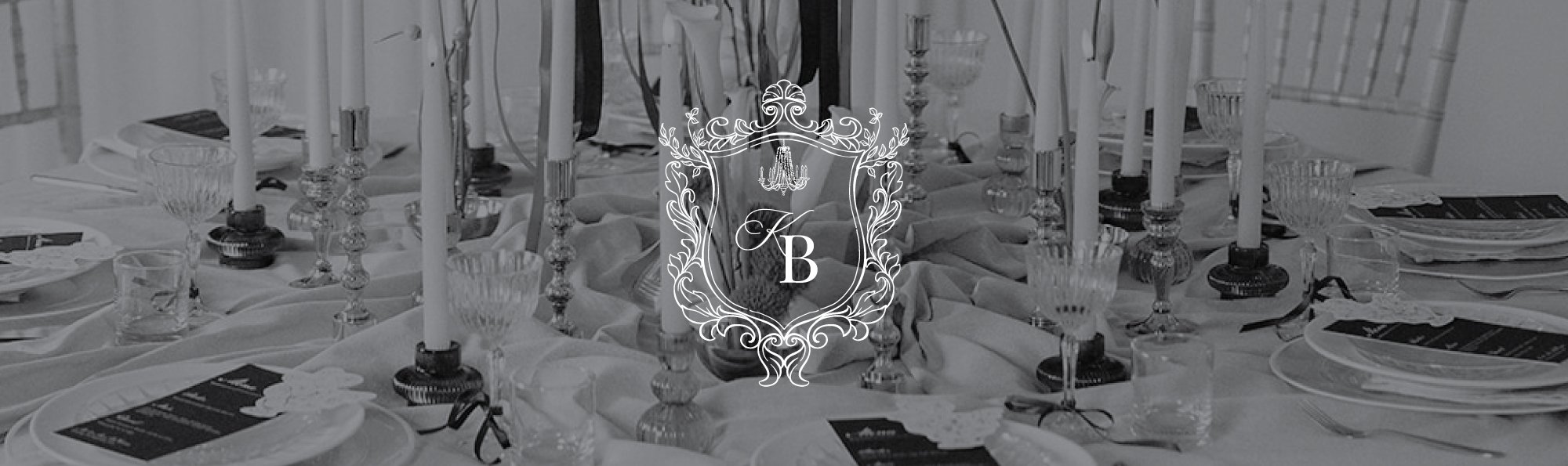 Elegant dining table decorated with white tablecloth, crystal glasses, and tall white candles in candlesticks, set for a formal event. A decorative emblem with the initials 'K B' and a chandelier design is overlaid in the center.