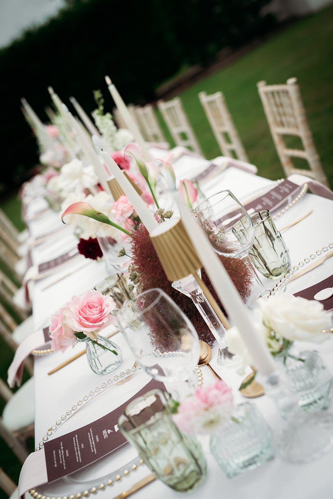 Elegant outdoor dining table decorated with pink and white flowers, candles, and glassware, set for a special occasion.