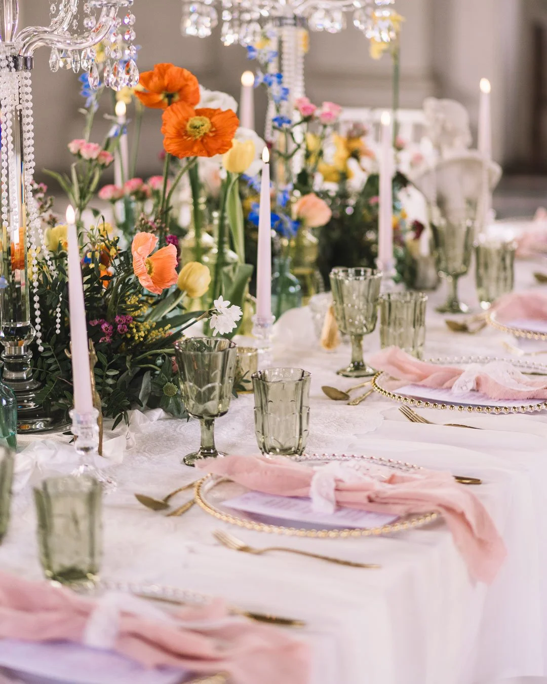 Elegant banquet table decorated with pink napkins, gold utensils, and glassware, with a floral centerpiece featuring colorful flowers and tall pink candles, set in a luxurious, well-lit room.