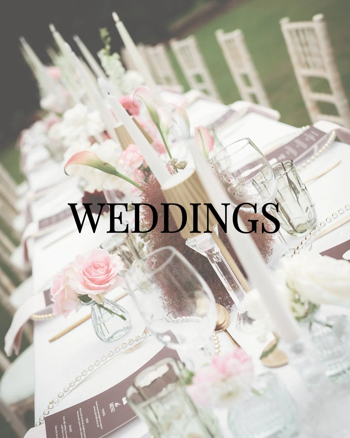 Elegant wedding reception table decor with pink and white flowers, candles, and glassware, set outdoors with chairs and greenery in the background.