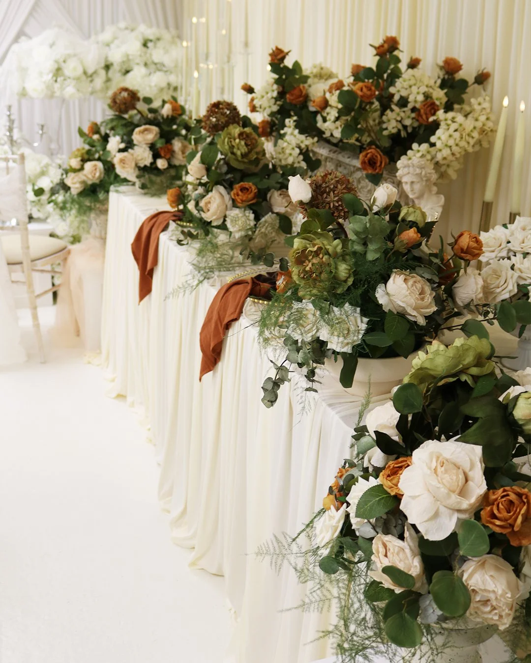 Long banquet table decorated with an arrangement of white, beige, and rust-colored flowers and greenery, draped with cream-colored fabric, in a decorated indoor setting with a cream-colored curtain backdrop.