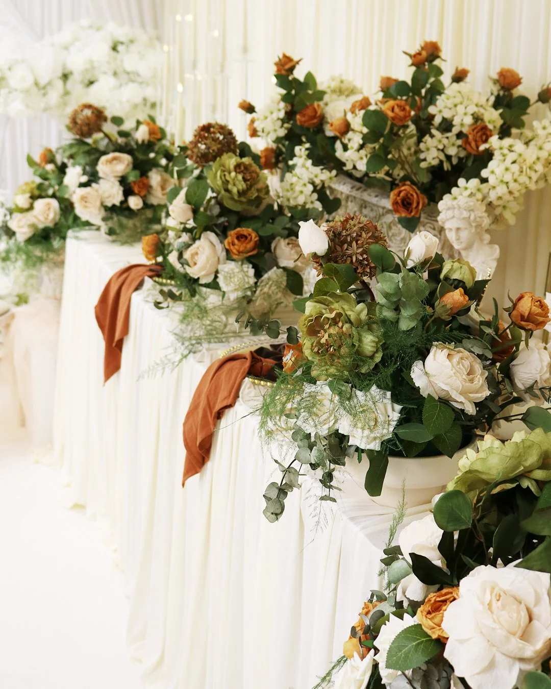 Elegant table decorated with floral arrangements of white, orange, and blush roses, green foliage, and dried flowers, with draped white fabric and rust-colored satin accents, set up for a formal event or wedding reception.