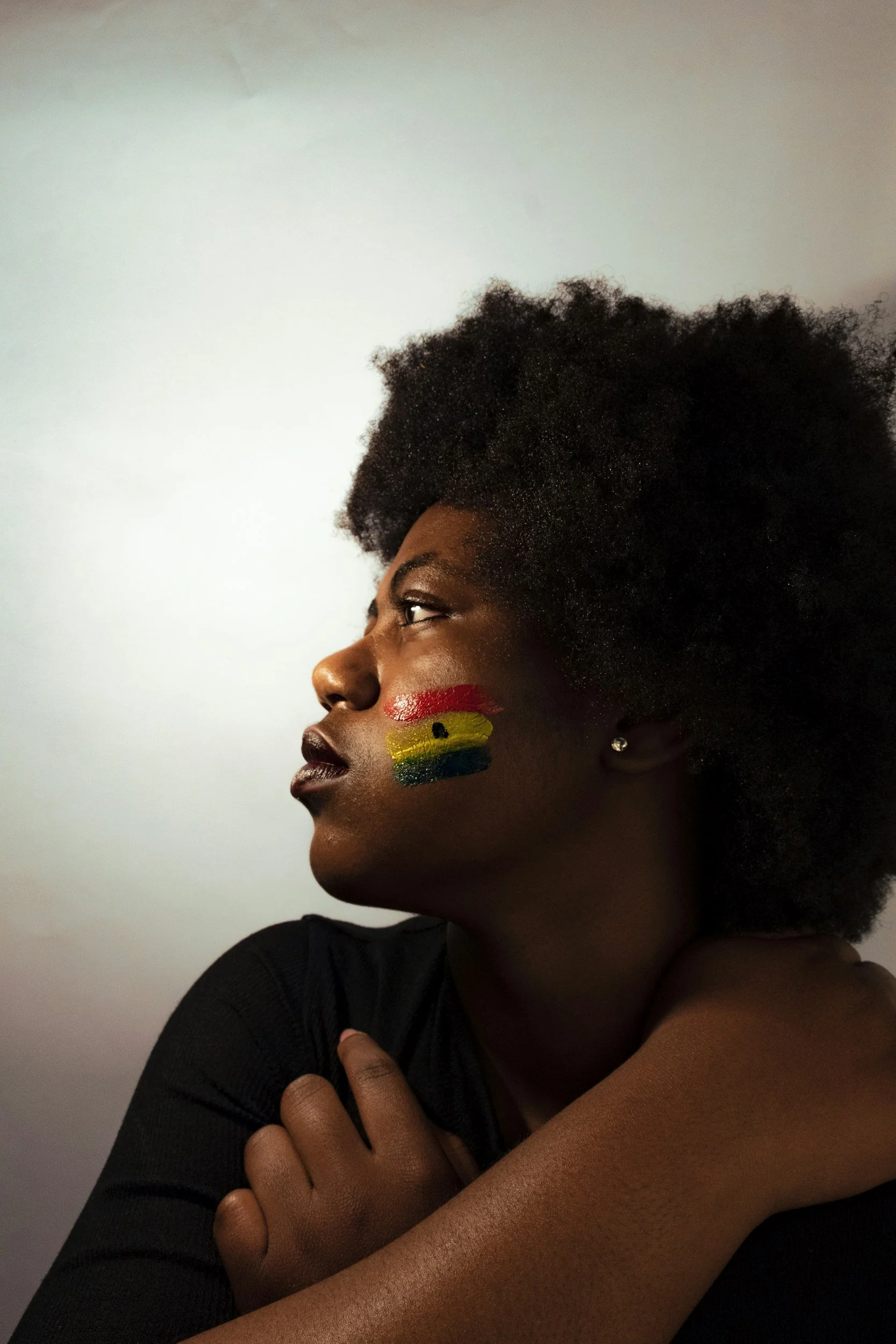 A woman with painted Ghanaian flag on her cheek, looking to the side, with natural hair and a black shirt, against a plain background.