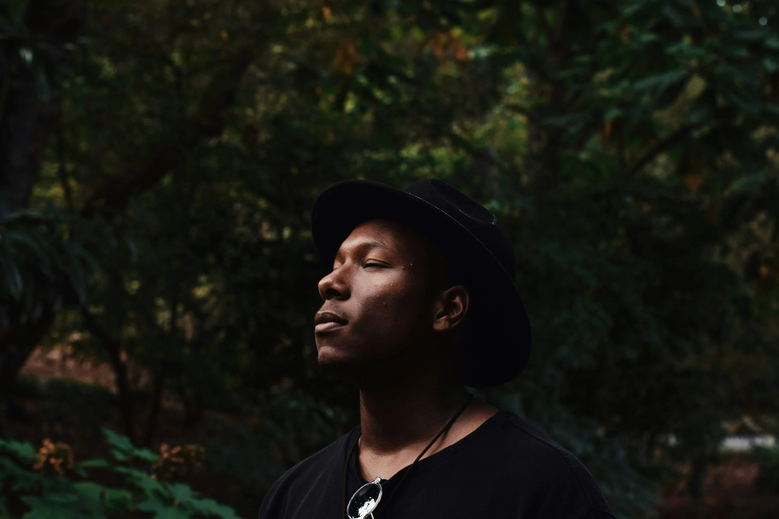 Person with dark skin wearing a black hat and black shirt, standing outdoors in a green, wooded area with sunlight filtering through the trees.