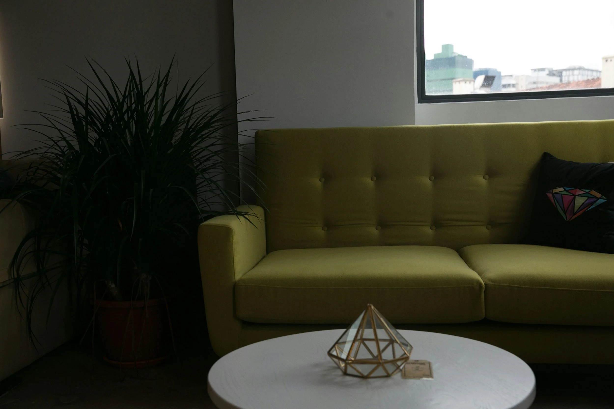 A green sofa next to a potted plant and a round white table with geometrical decor, in front of a window with a city view.