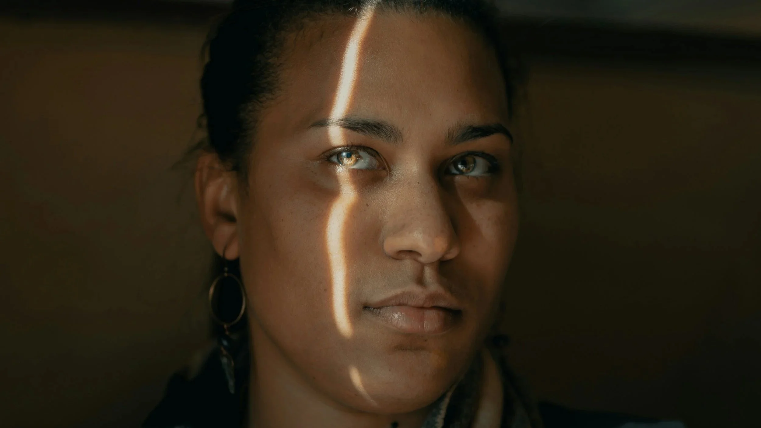A close-up of a woman with blue eyes, dark hair, wearing earrings, with a light stripe crossing her face, in a dimly lit environment.