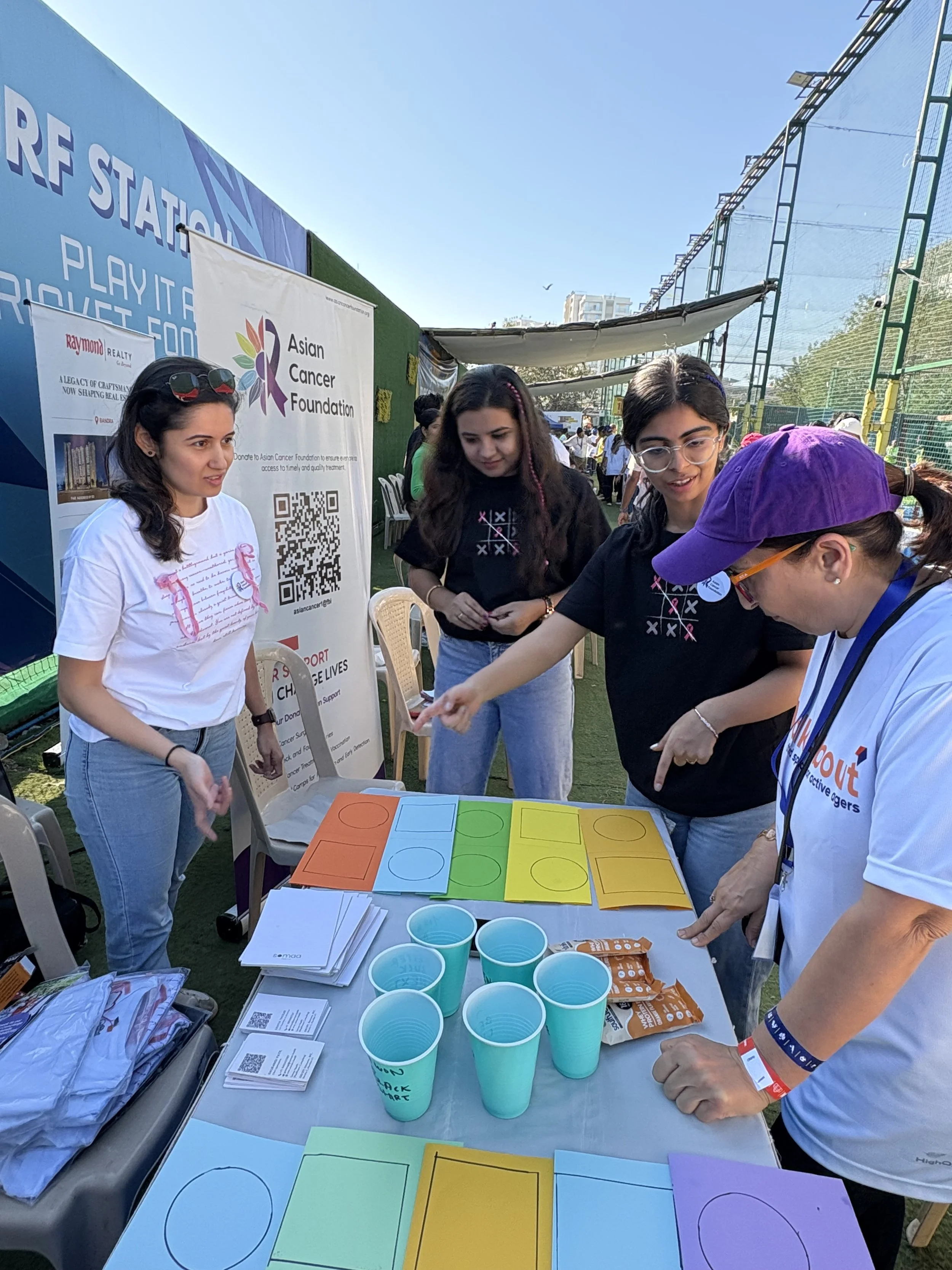 Group of people gathered around a table at an outdoor event with colorful paper sheets, cups, flyers, and a banner promoting the Asian Cancer Foundation.