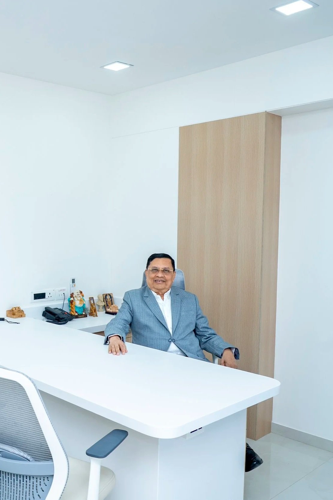 Dr Kulin Kothari in a gray suit sitting at a white office desk in a modern, minimalist office with a wooden feature wall, a white chair, and decorative statues on the desk.