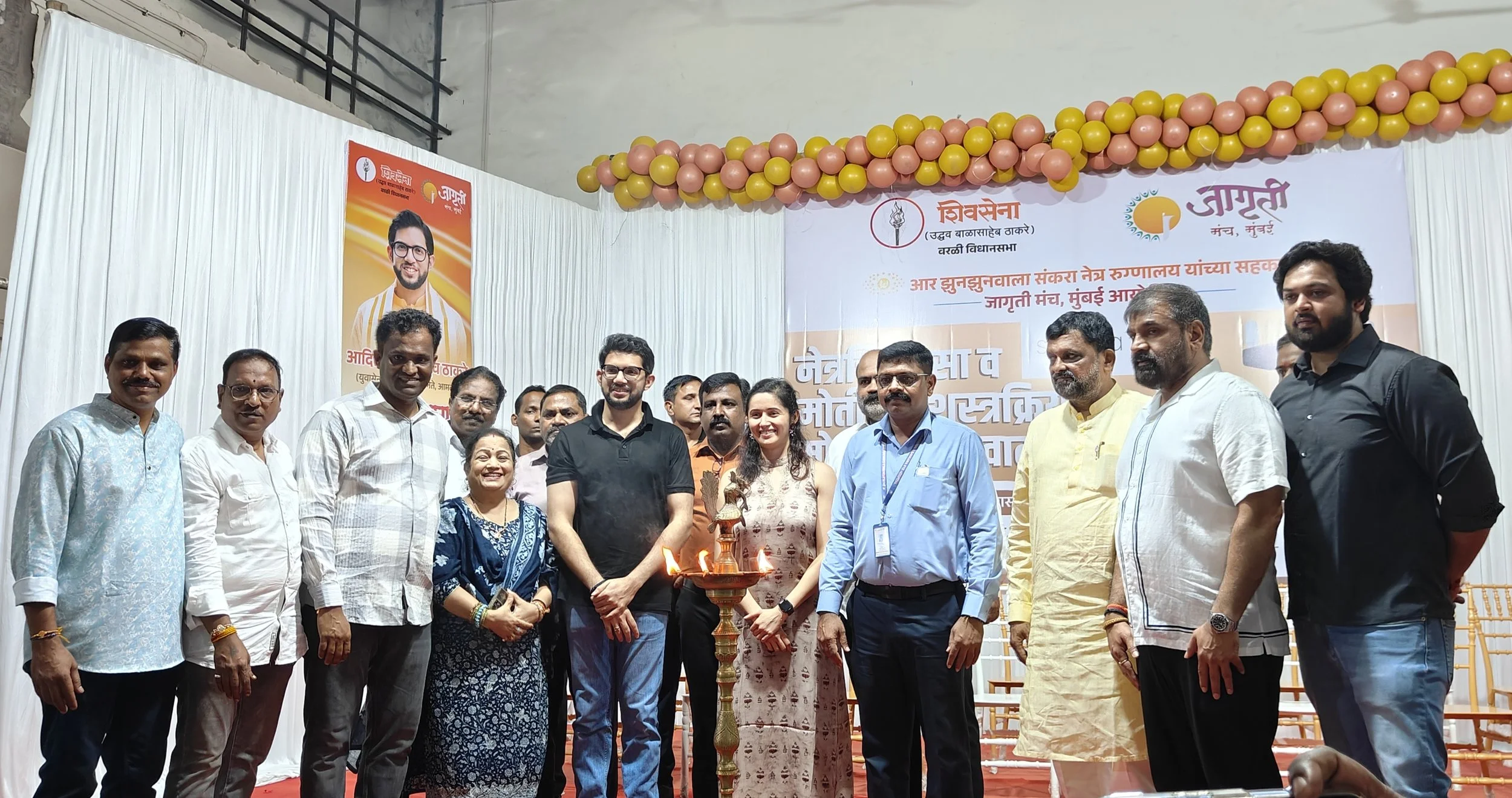 A group of people gathered at an event with a banner and balloons in the background. They are standing around a traditional Indian lamp, which is lit, indicating a ceremonial or festive occasion.