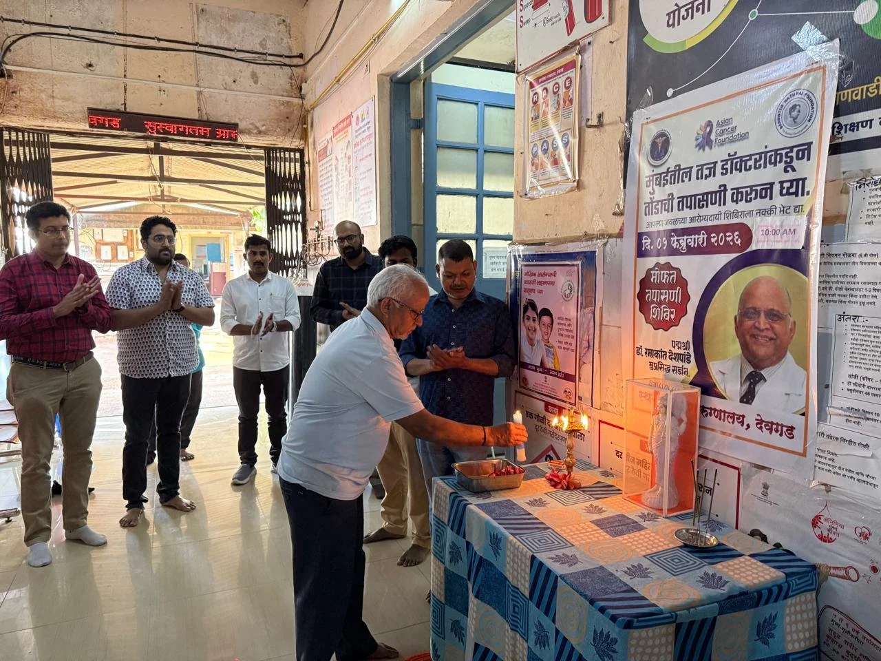 A group of men participating in a prayer or ritual indoors, lighting a lamp on a decorated table with banners and posters in the background.