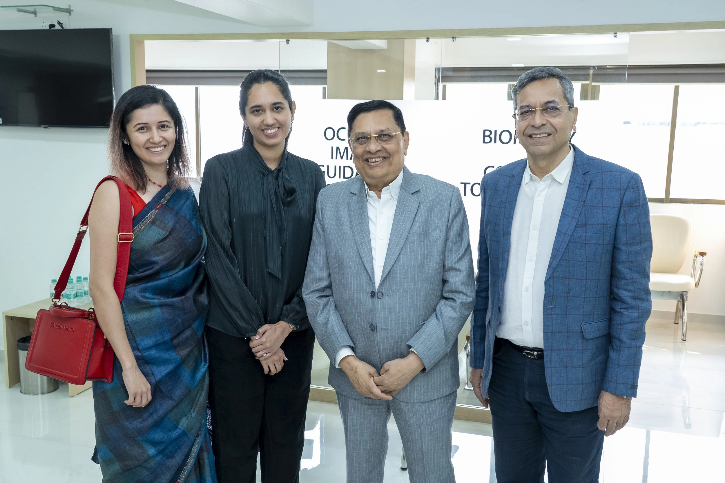 Five professionally dressed people posing indoors, smiling at the camera. The background has a whiteboard with partial text and a modern office setting.